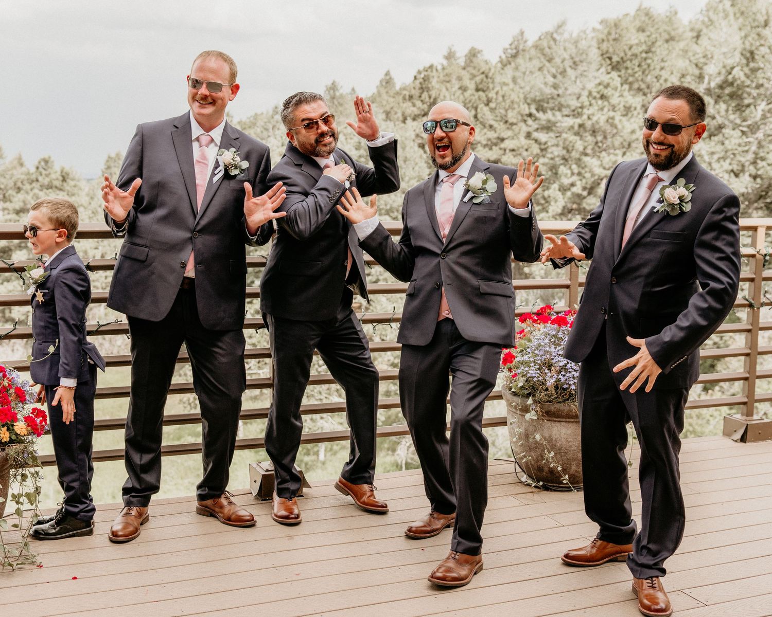 Group of groomsmen in gray suits and brown shoes having fun posing on a wooden deck outdoors.