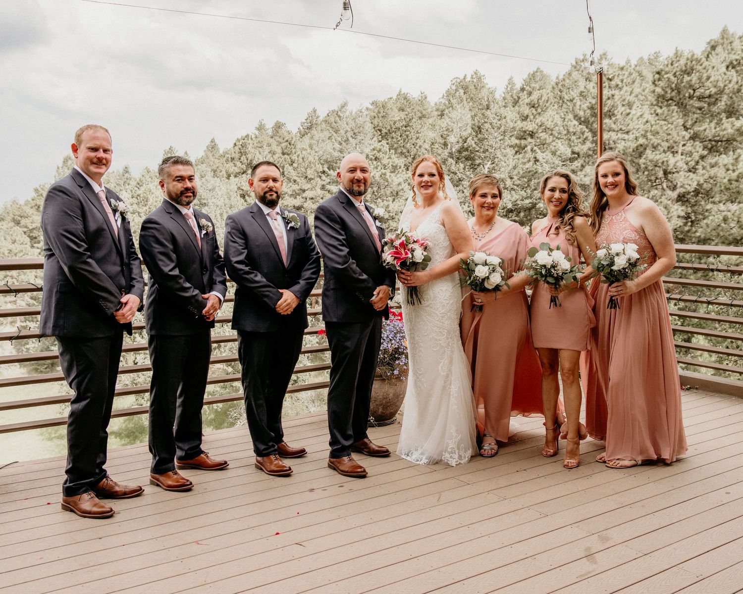 Wedding party in formal attire stands on wooden deck with forest backdrop during outdoor ceremony celebration.