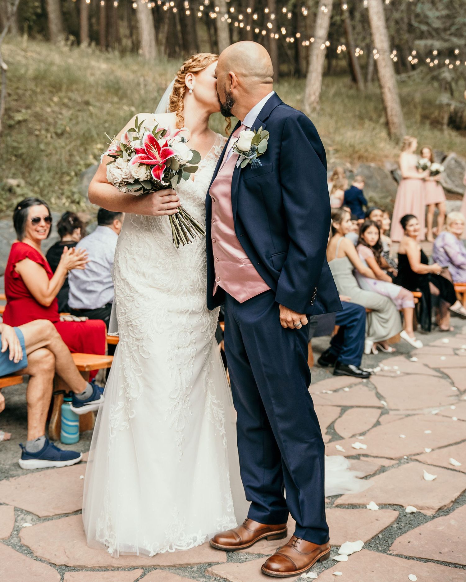 A romantic outdoor wedding ceremony with string lights overhead as a couple shares a kiss on rustic stone paving.