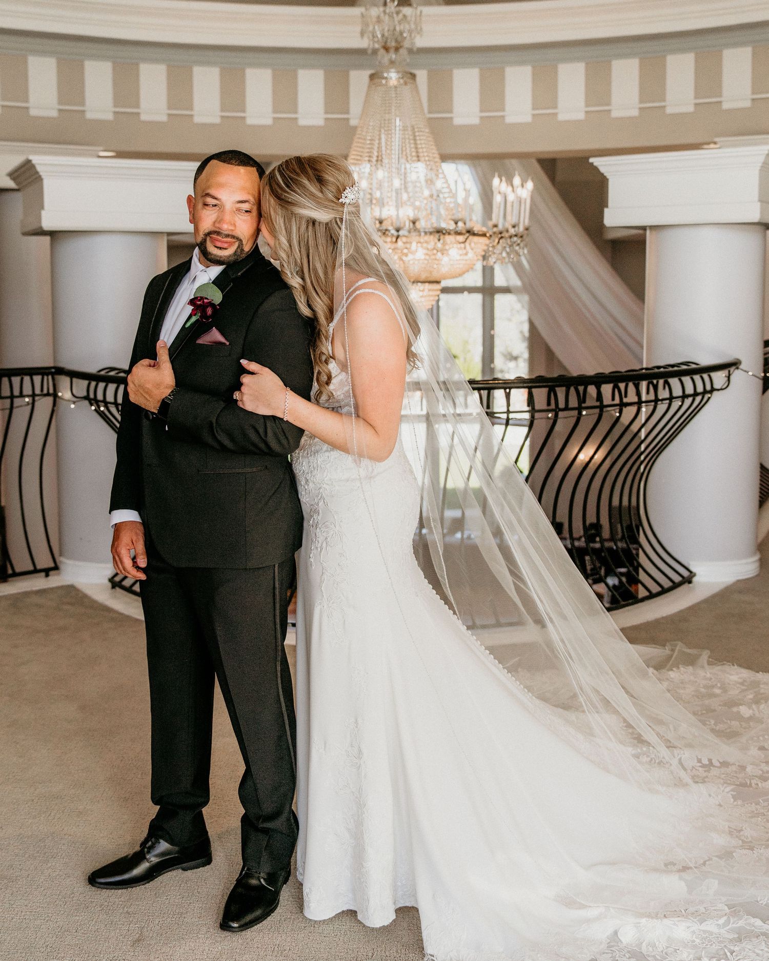 A couple shares a joyful moment during their wedding in an elegant venue with a crystal chandelier and curved staircase.