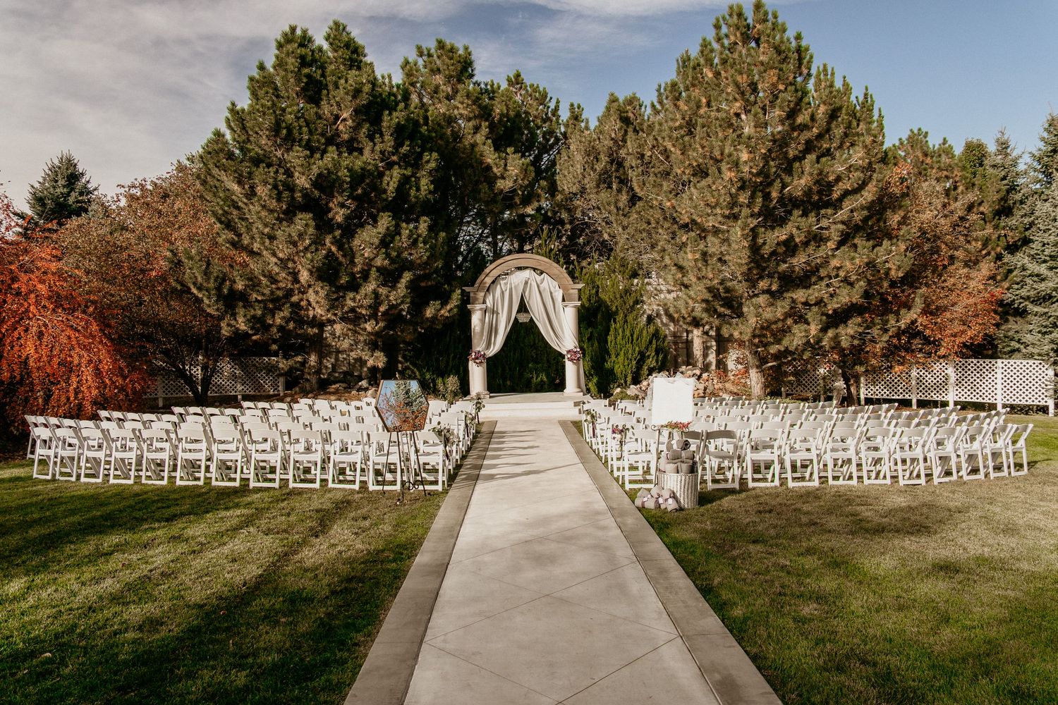 A decorated wedding arch with white chairs arranged outdoors on a lawn surrounded by autumn pine trees.