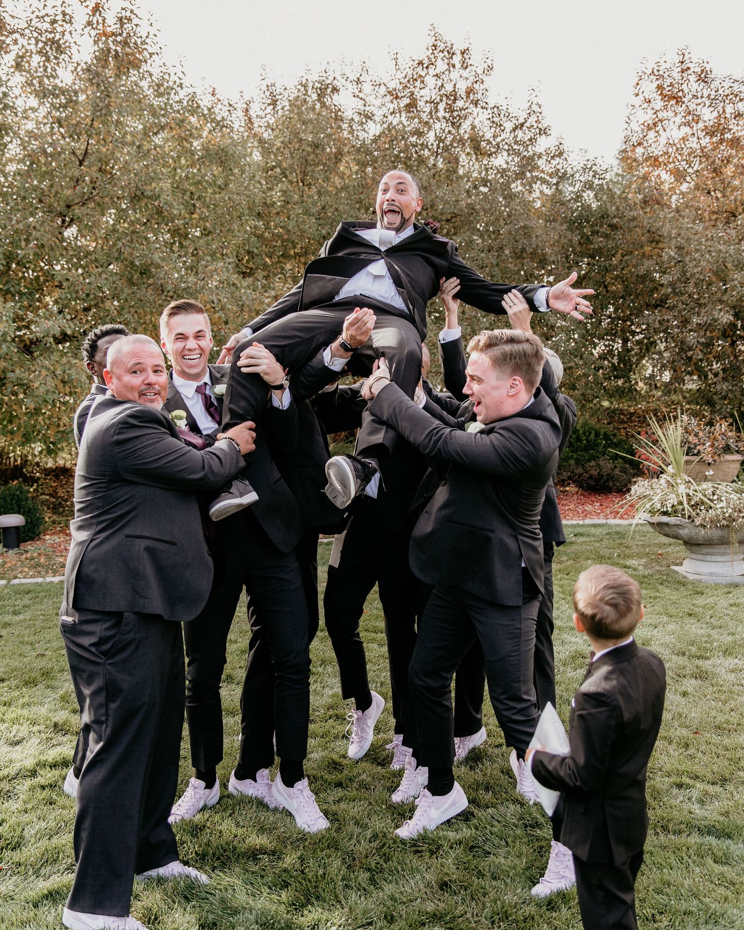 A group in formal black suits and white sneakers lift someone up in celebration outdoors during a special occasion.