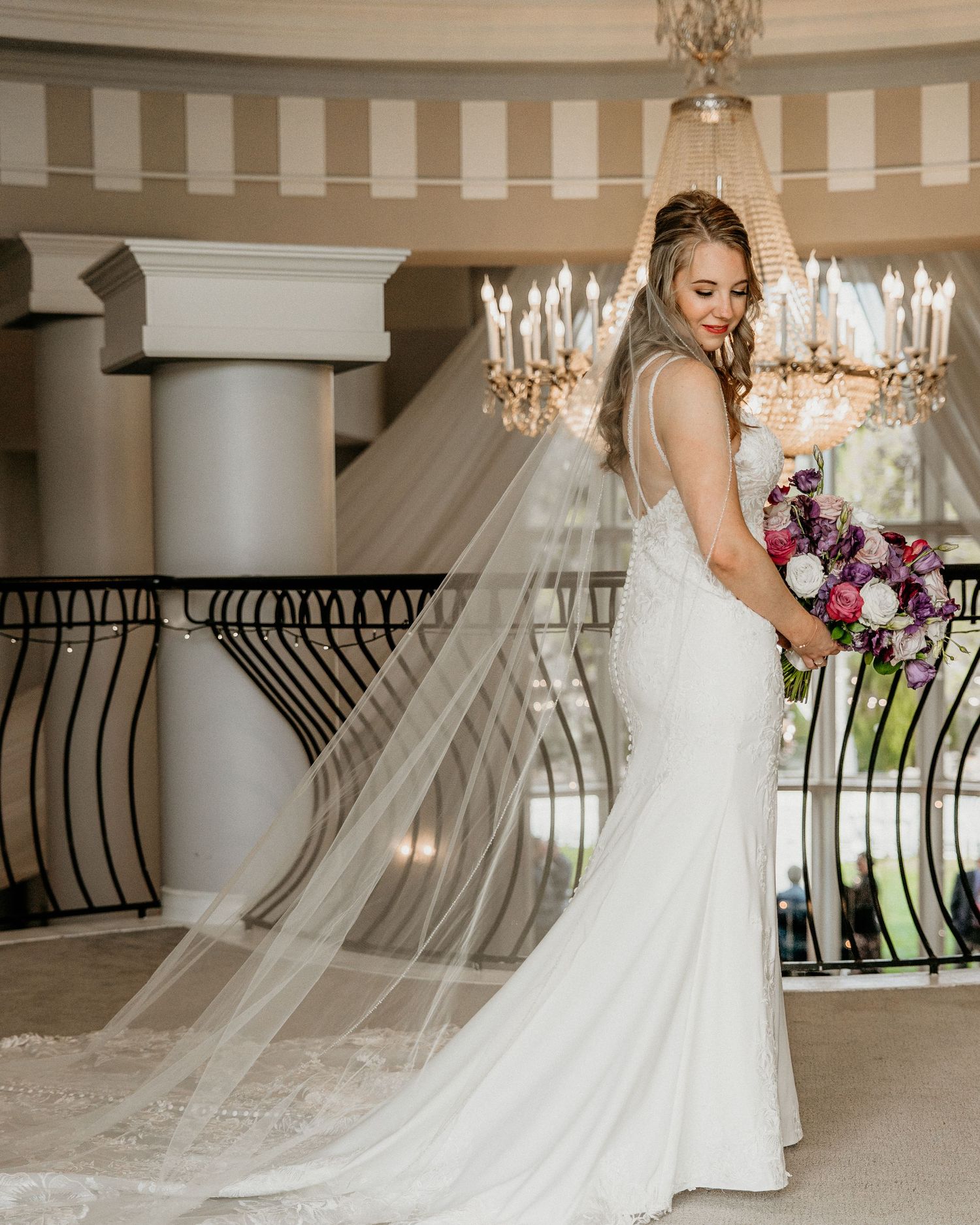 Bride in elegant fitted lace wedding gown and long veil poses beside ornate railing under crystal chandelier.