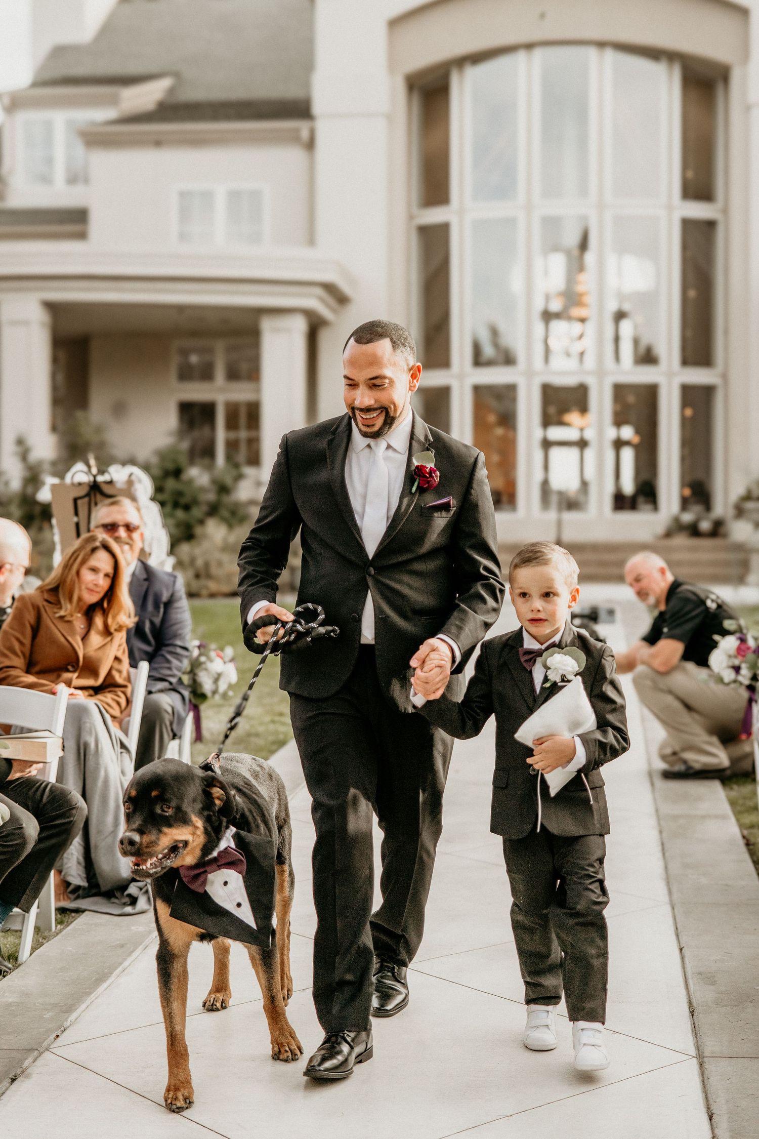 A joyful wedding moment as a suited figure walks down the path with a young child and a dog in formal attire.