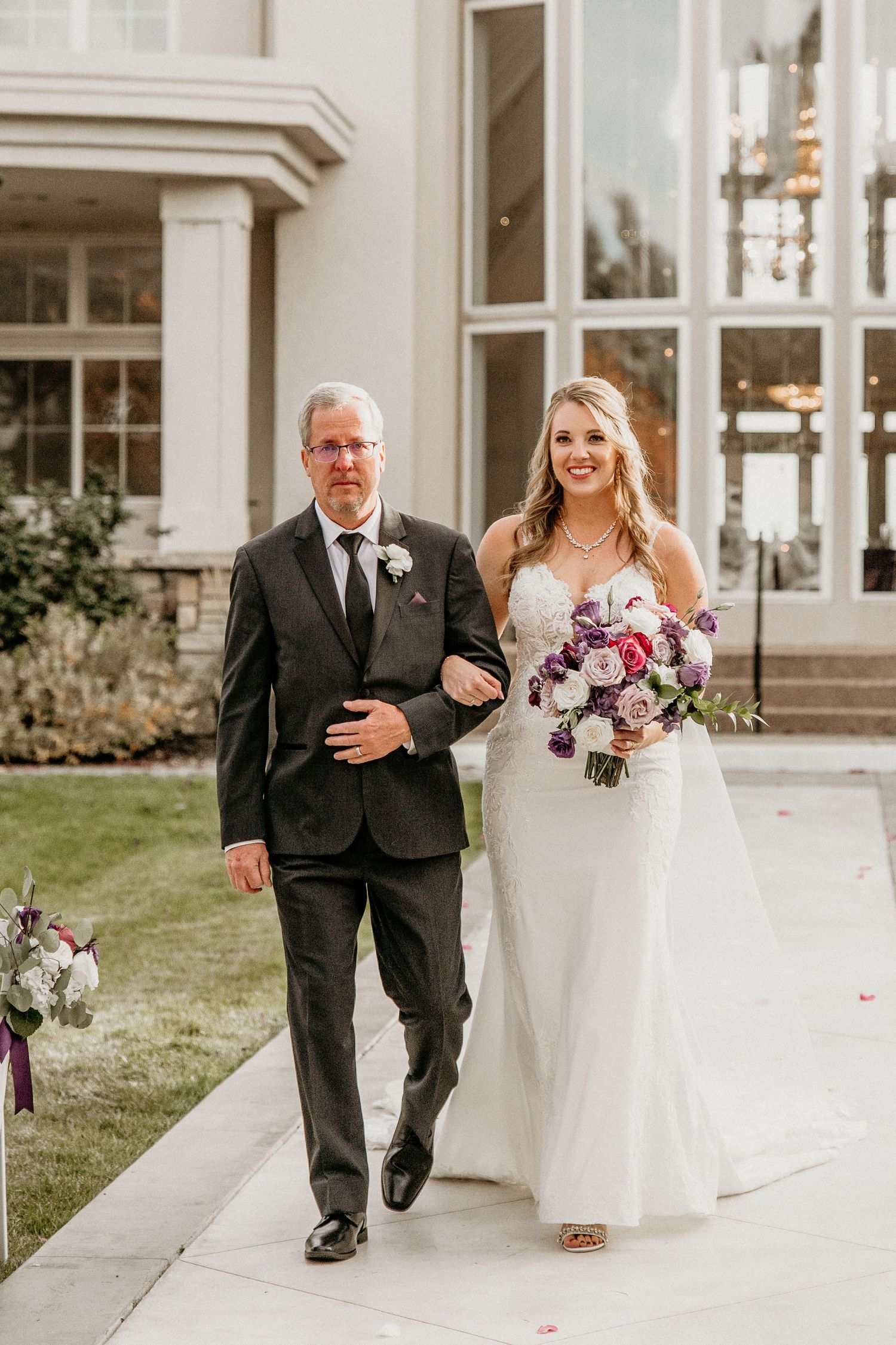 Two people walk arm in arm at an elegant wedding venue with a purple floral bouquet and white gown.