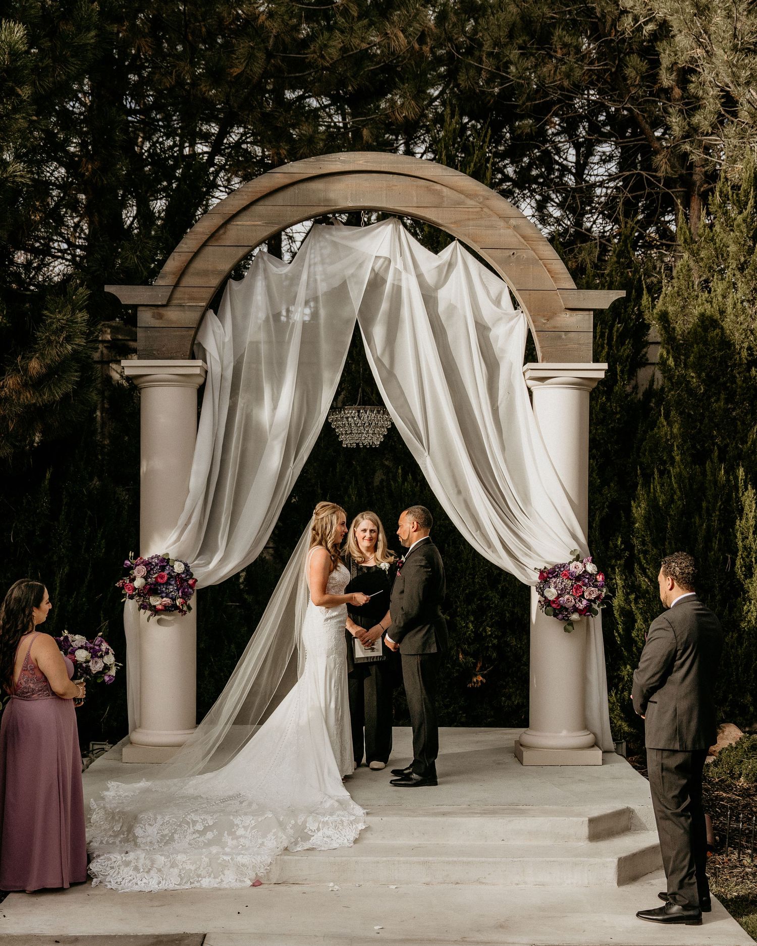 Wedding ceremony taking place under an elegant stone arch draped with white fabric and floral arrangements.