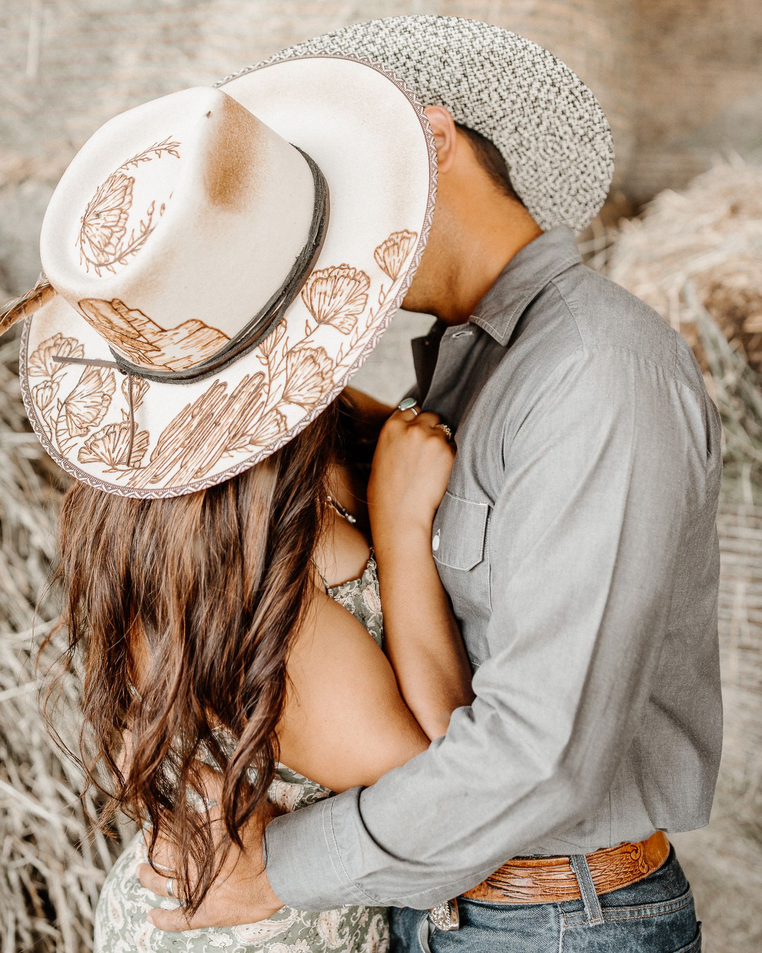 Romantic couple embraces in a field, with woman wearing decorative white western hat and wavy brown hair.