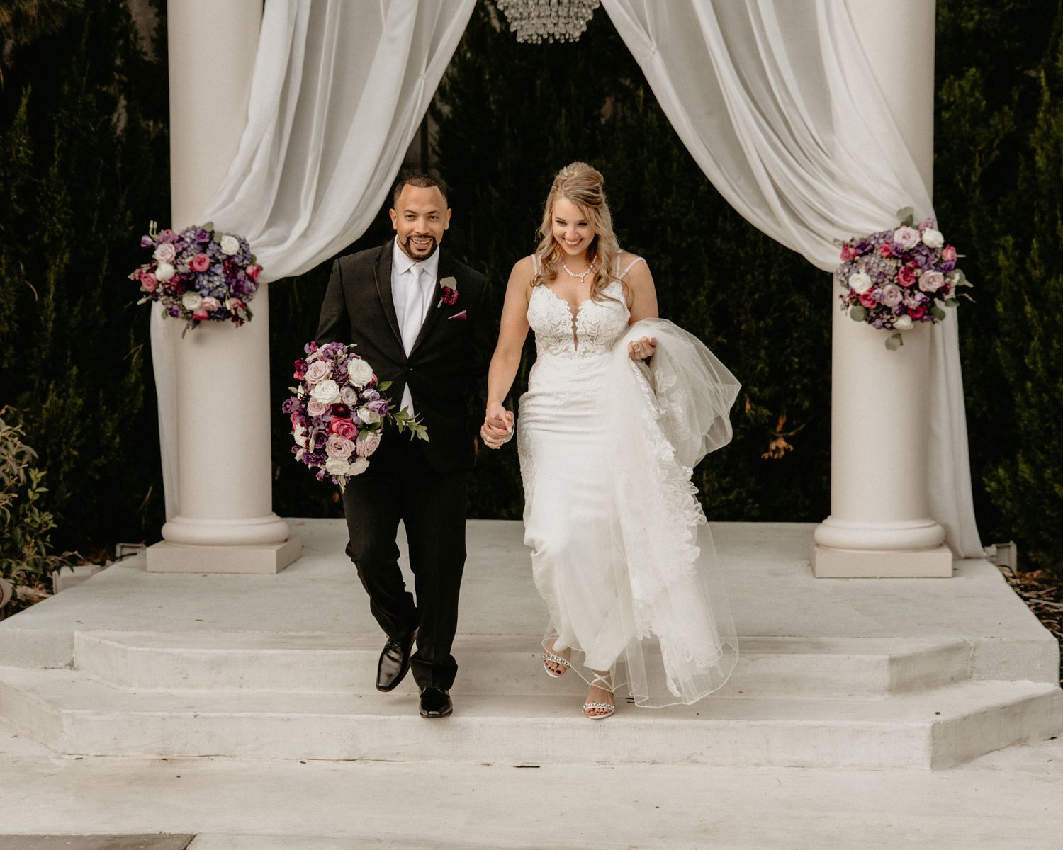 Happy newlyweds exit white ceremony arch with pink floral arrangements during outdoor wedding celebration.