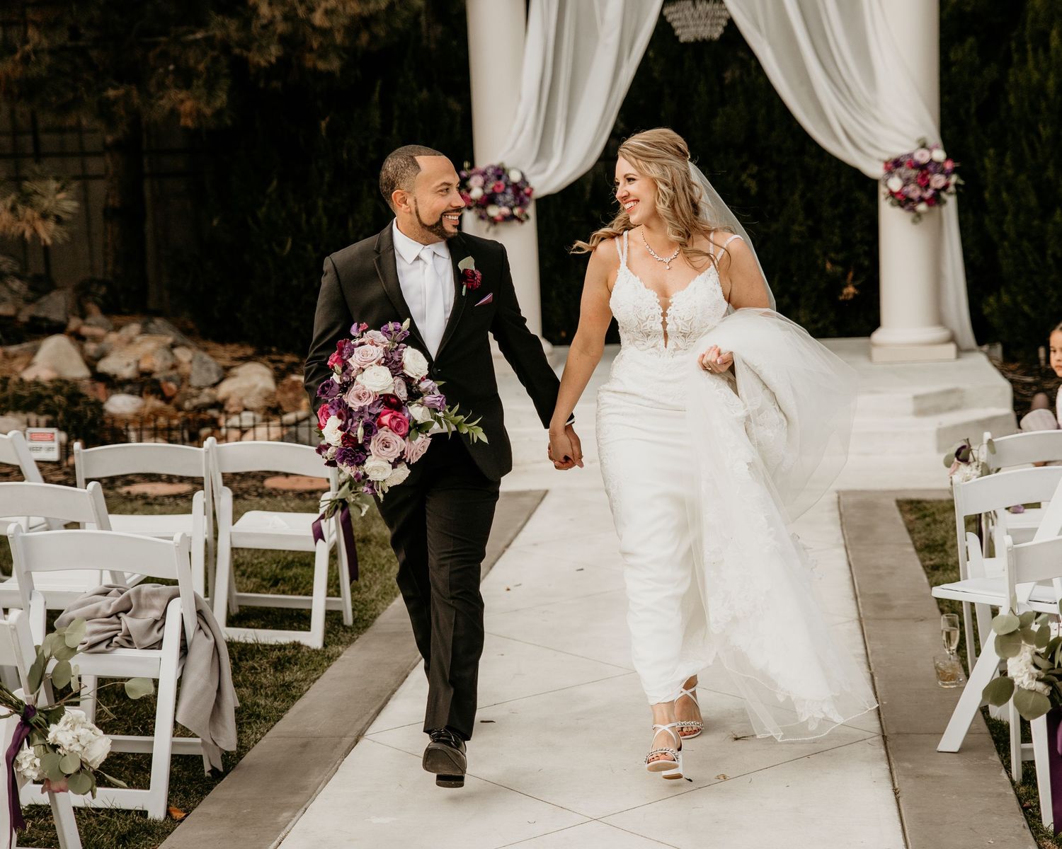A joyful couple walking down the aisle together after their outdoor wedding ceremony with white decor and purple flowers.