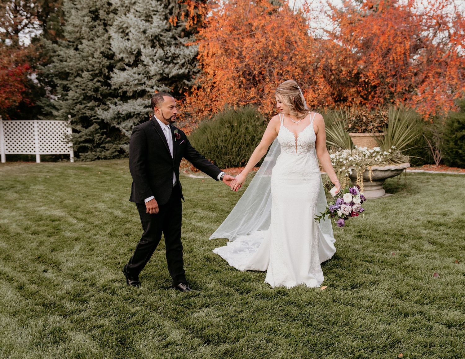 A couple in wedding attire walks hand in hand on a green lawn with autumn foliage and evergreens in the background.