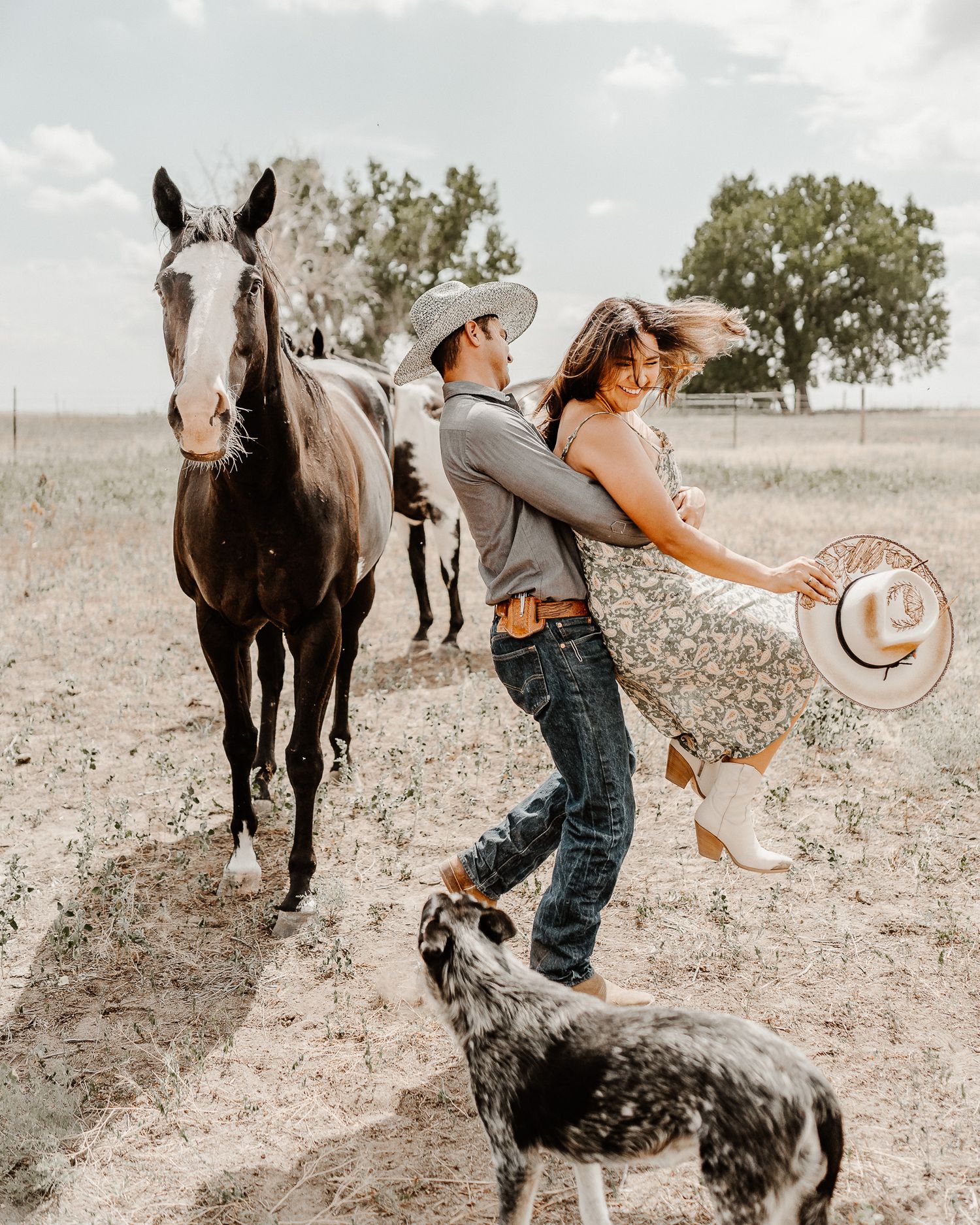 A playful western-themed couple dances in a field with their horse and spotted dog on a sunny day.