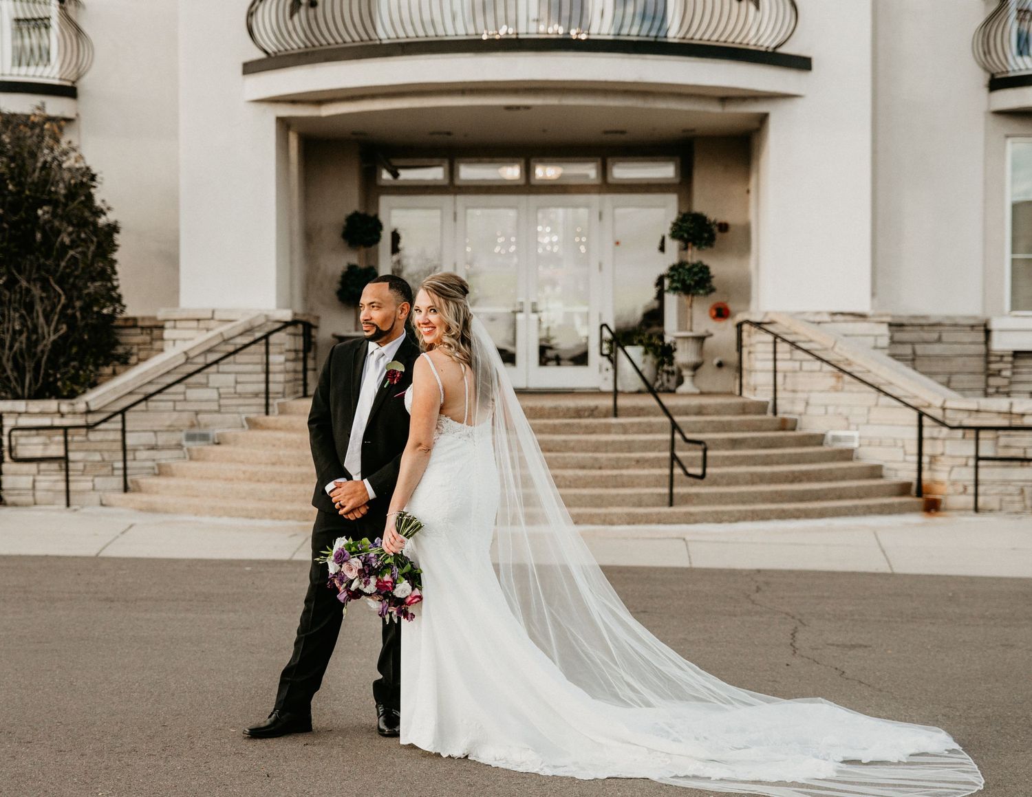 A bride in a flowing white gown with a veil stands next to a groom in a tuxedo at the entrance of an elegant venue.