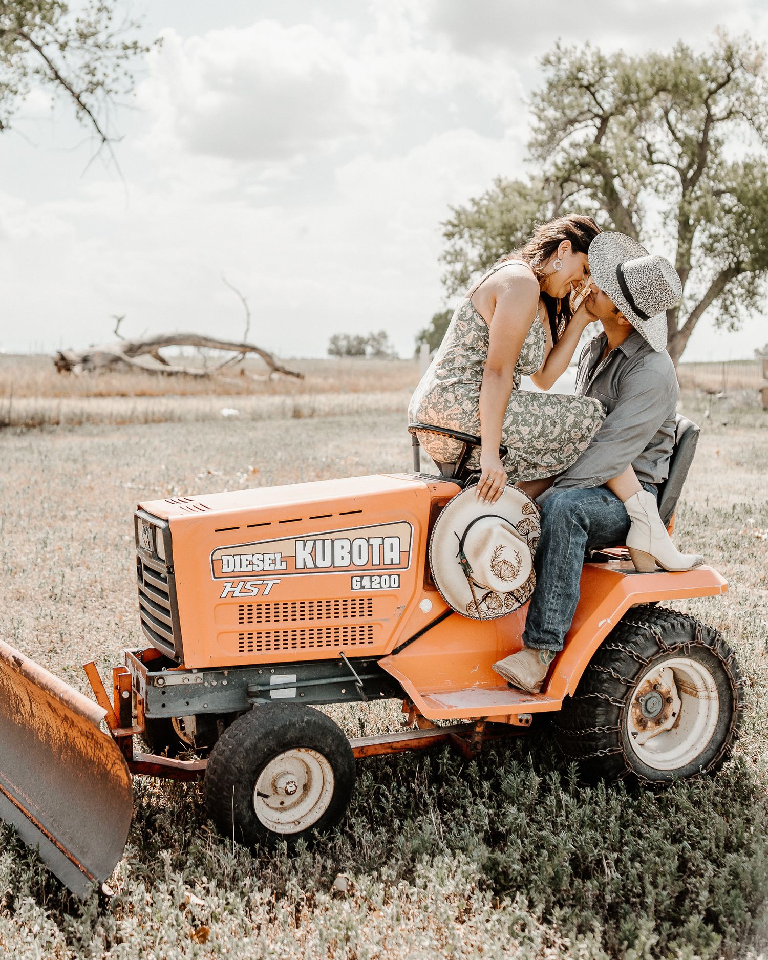 A couple shares a romantic moment on an orange Kubota tractor in a rustic country field with trees in the background.