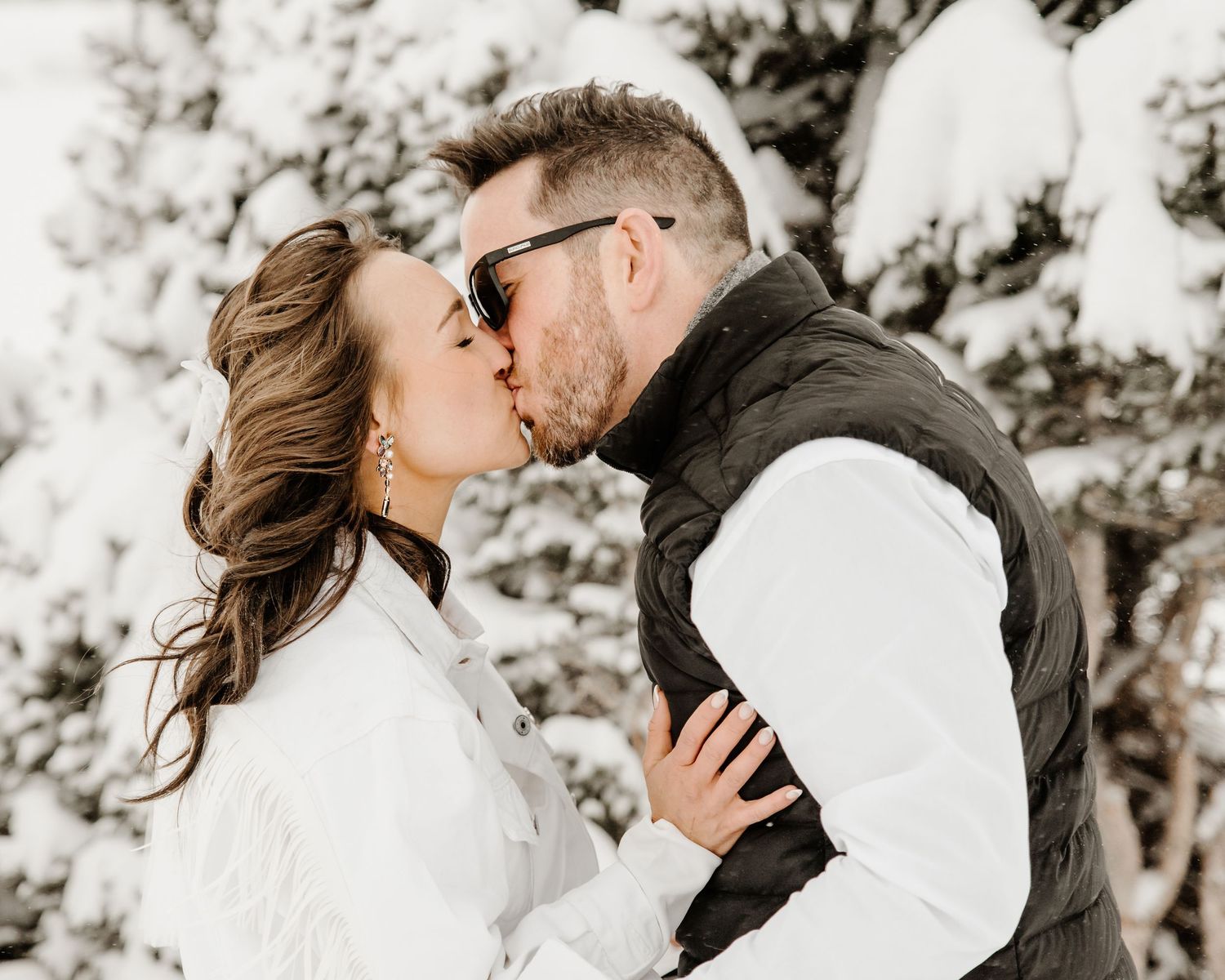 A couple in white and black winter attire share a romantic kiss in front of snowy evergreen trees.