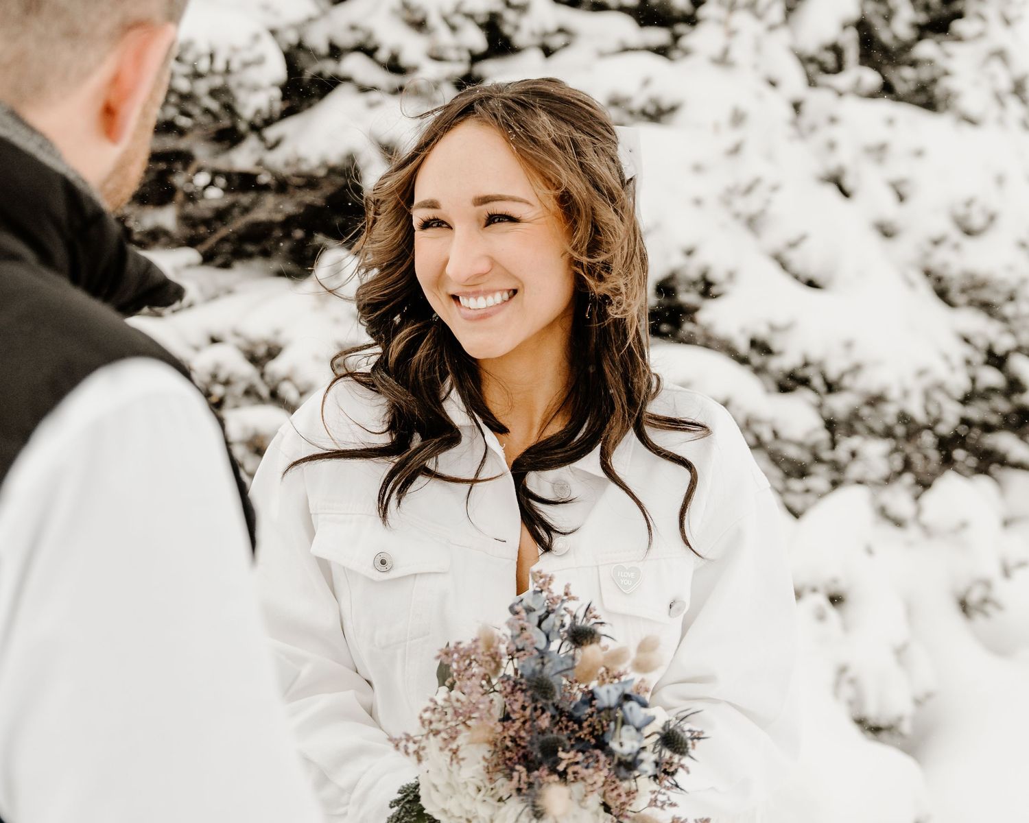 Winter wedding scene with a person in a white jacket holding a muted blue and brown floral bouquet in snowy setting.