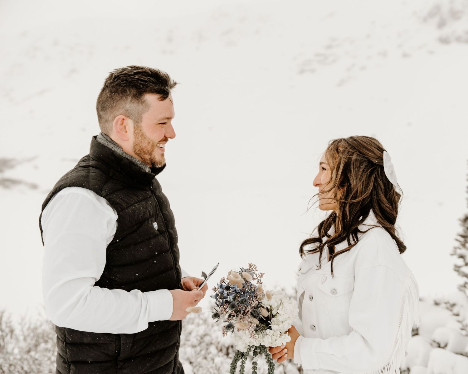 A couple shares a tender moment during their winter wedding ceremony while holding a bouquet in the snowy landscape.