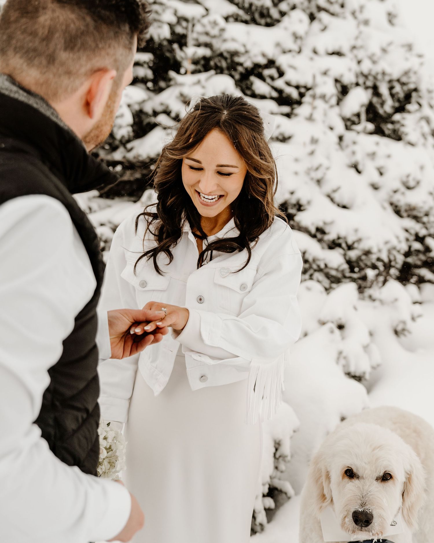 A couple shares a joyful winter engagement moment in the snow with their white dog beside them.