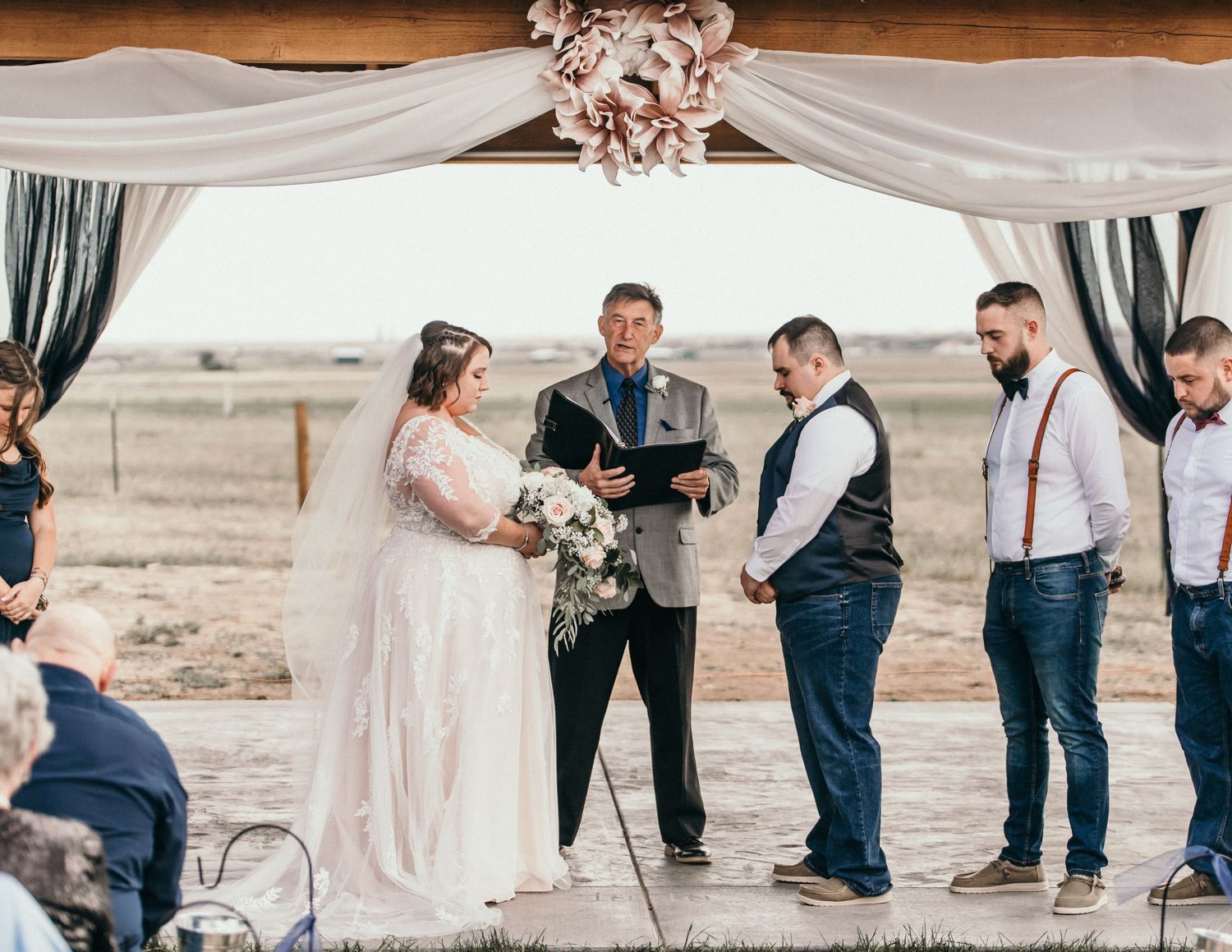 A wedding ceremony takes place under a draped arch in a rustic outdoor setting with a small wedding party.