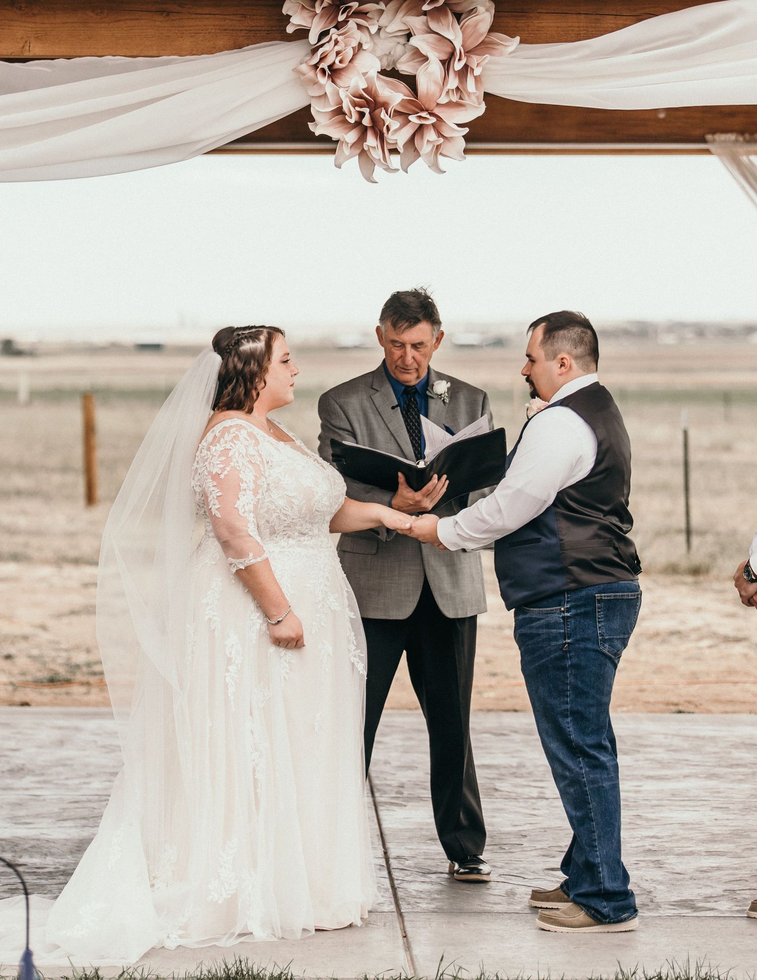 Wedding ceremony taking place outdoors under a wooden arch with white fabric draping and floral decor.
