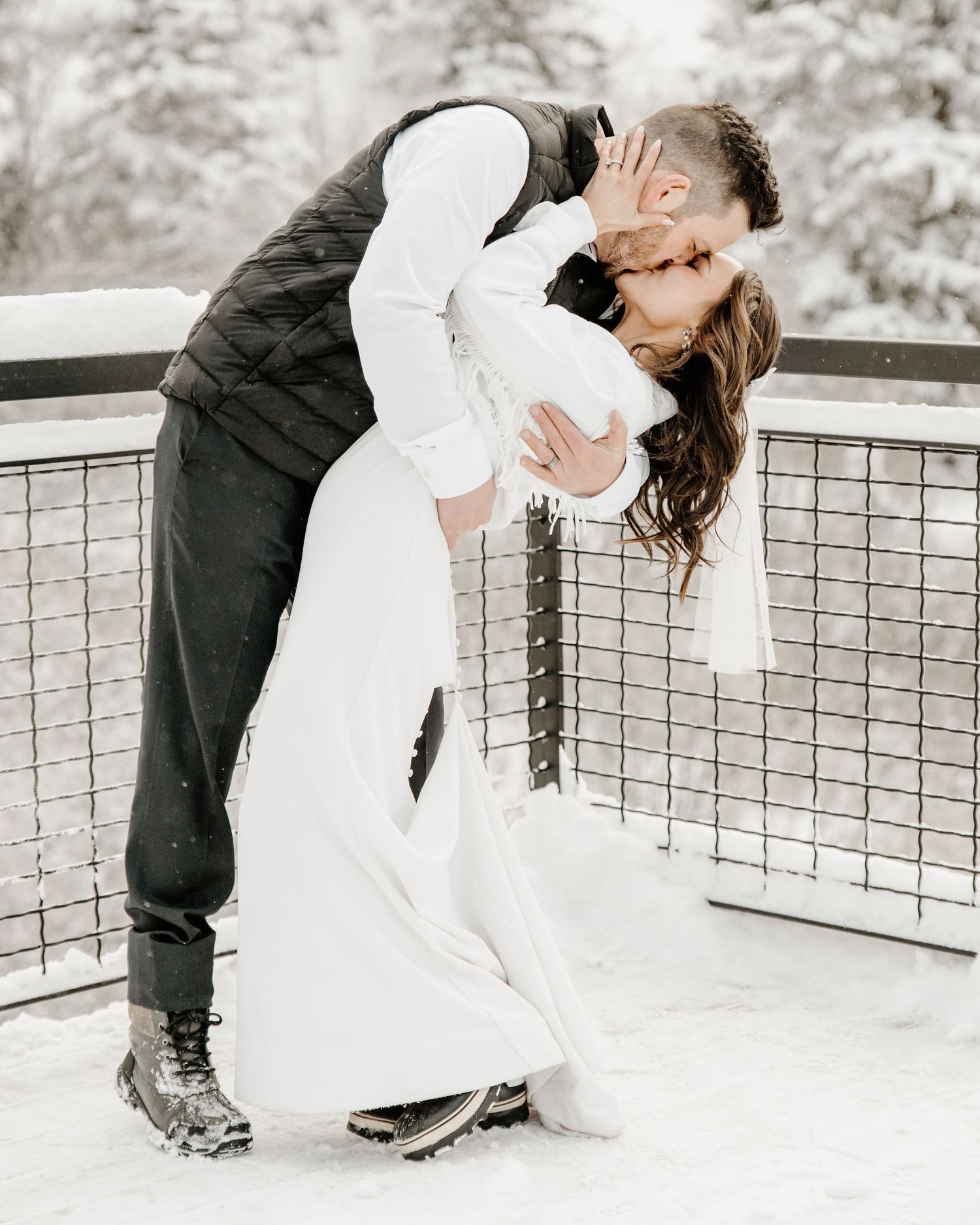 A romantic winter wedding portrait of a couple in classic white dress and dark vest embracing on a snowy outdoor deck.