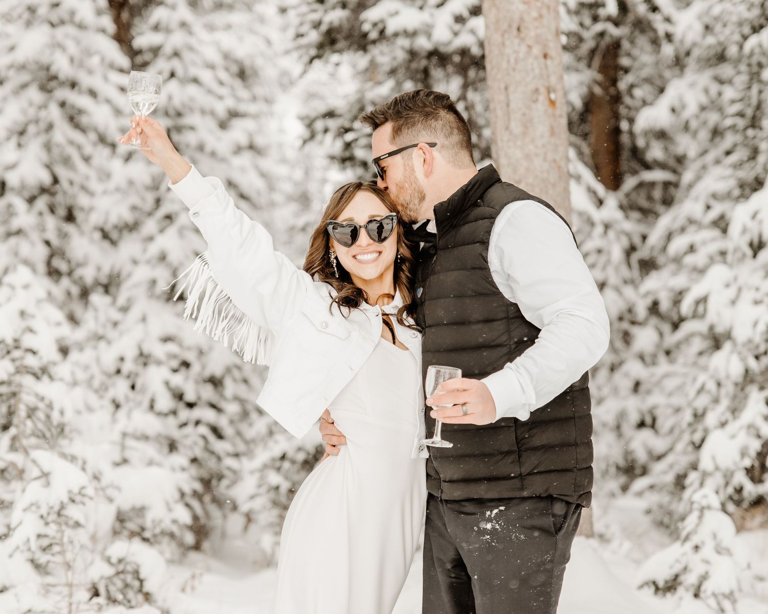 Couple embraces in a snowy forest setting with white winter wonderland backdrop while one person raises their arm in celebration.