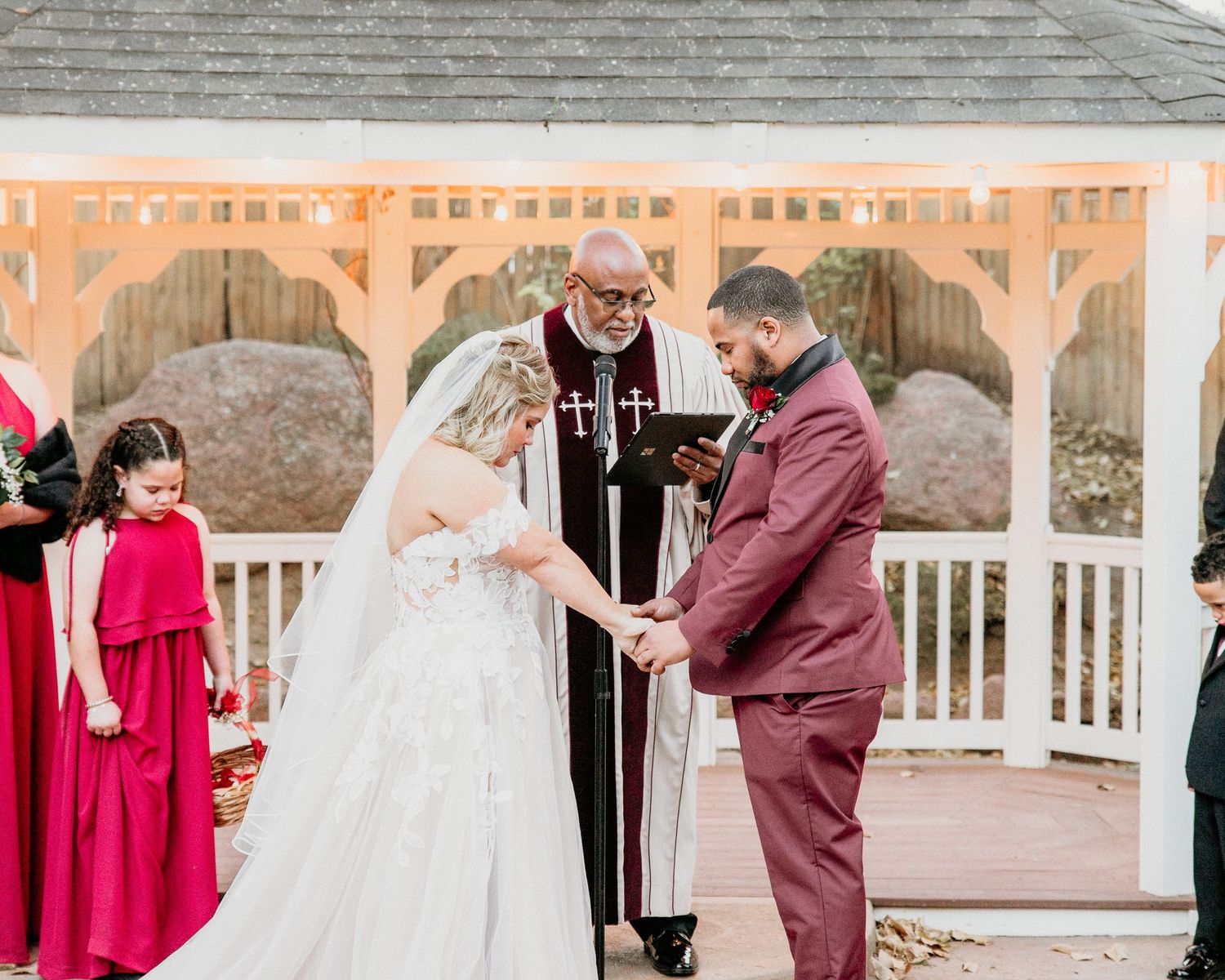 Wedding ceremony taking place at a white wooden gazebo with burgundy accents and fall decor.