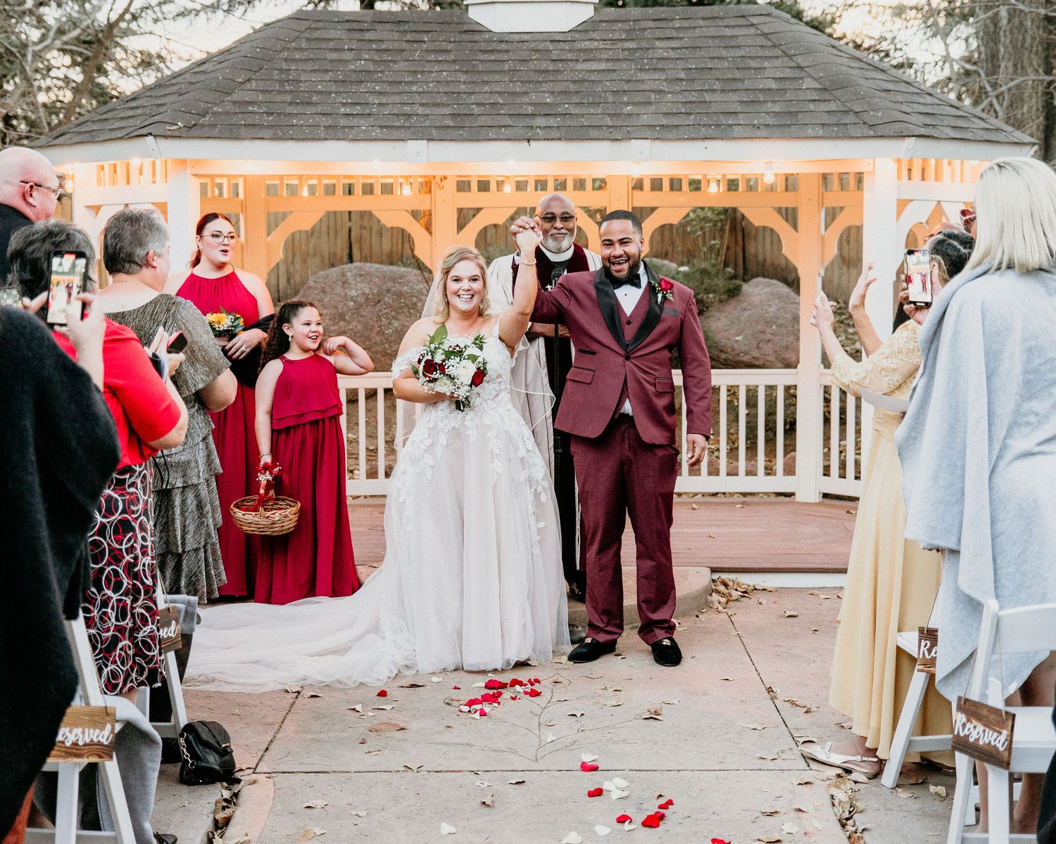 A wedding ceremony takes place at a white gazebo decorated with rustic wooden beams and red rose petals.