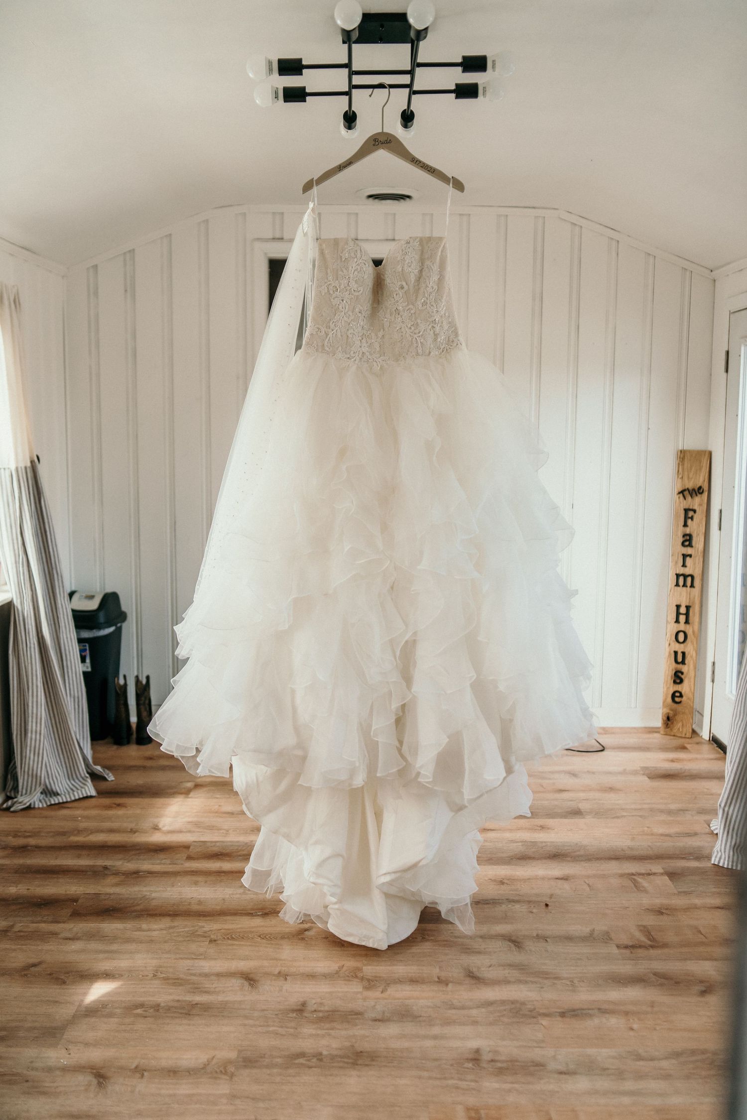 White layered wedding dress hanging on display in a rustic room with wooden floors and white walls.