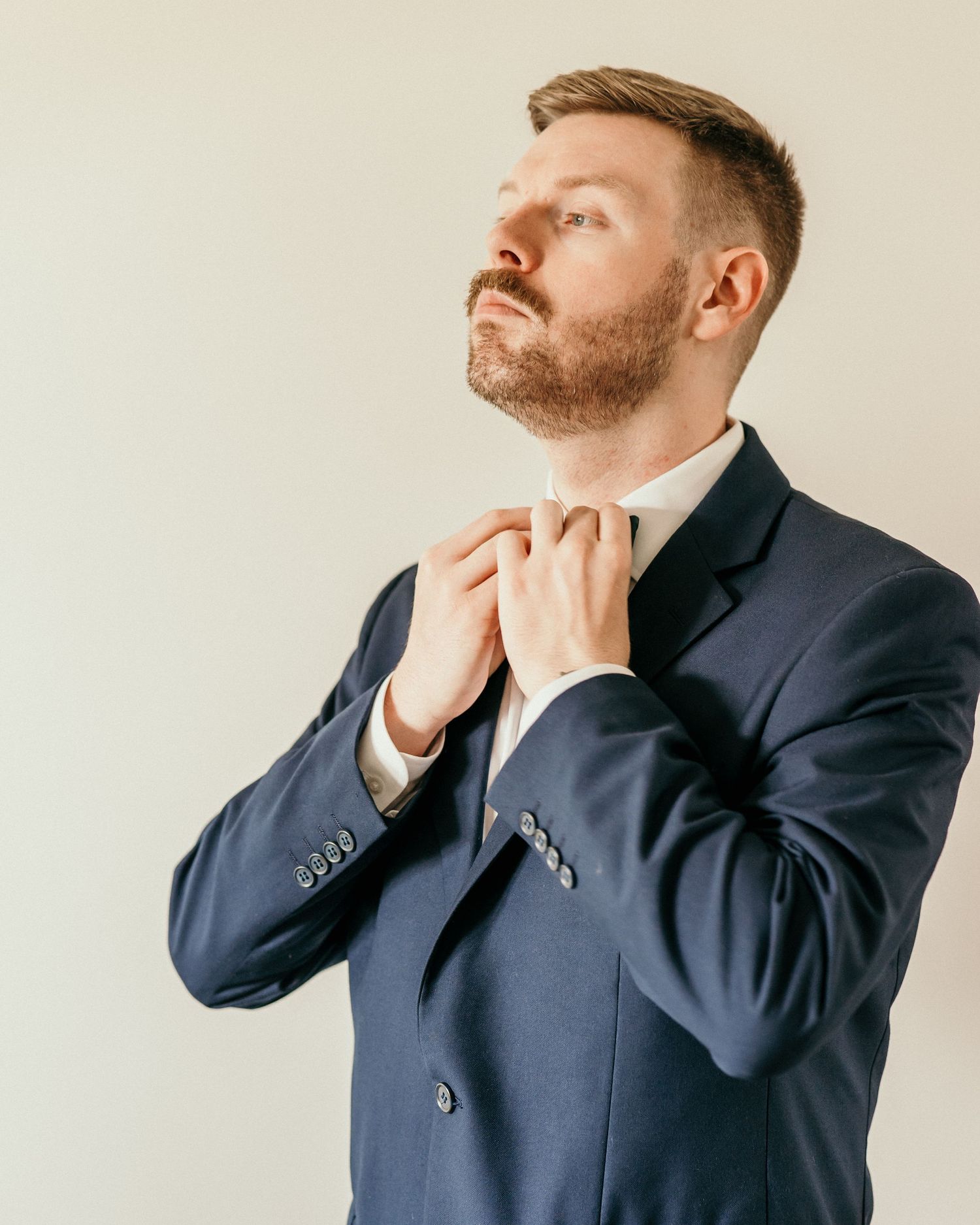 Professional in navy suit adjusts collar while looking upward against neutral background.
