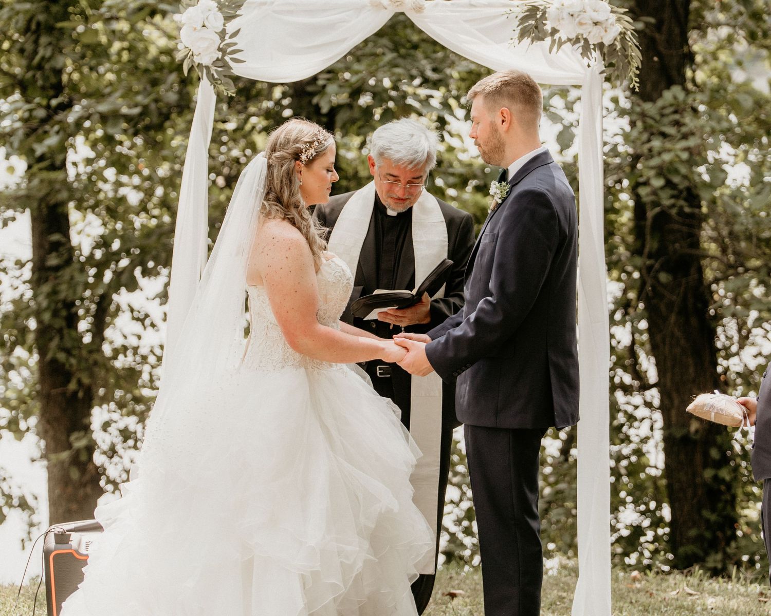 A couple exchanges vows during an outdoor wedding ceremony beneath a white fabric arch in a wooded setting.
