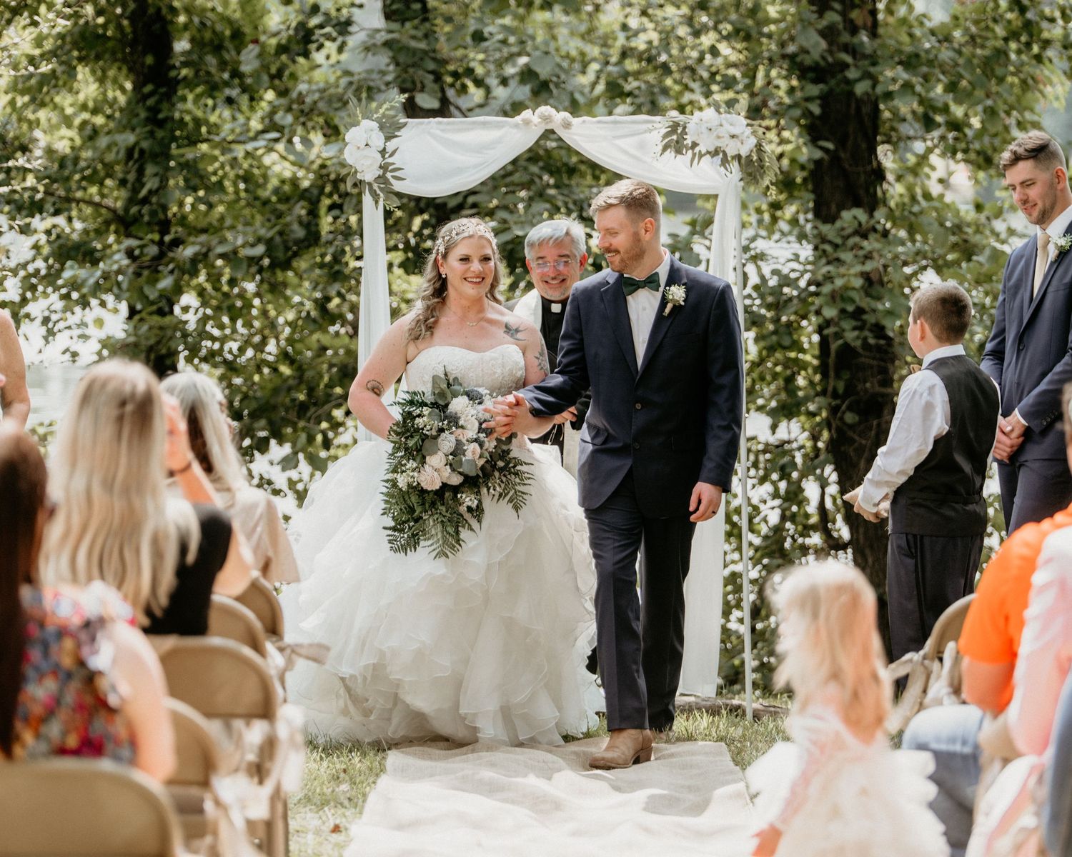 Couple walks down aisle at outdoor garden wedding ceremony under white arch surrounded by guests and greenery.