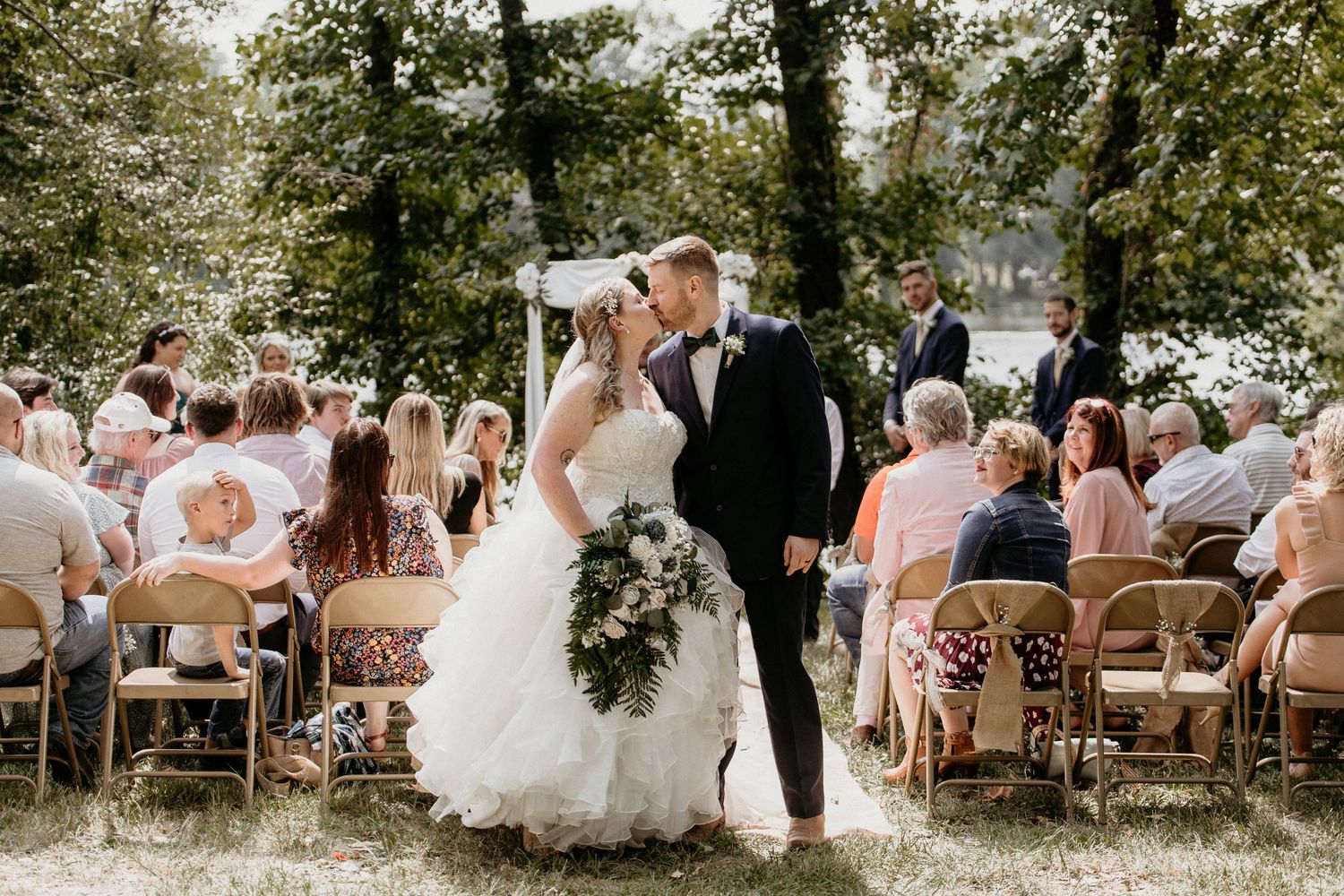 A couple shares their first kiss as newlyweds during an outdoor wedding ceremony surrounded by guests on wooden chairs.