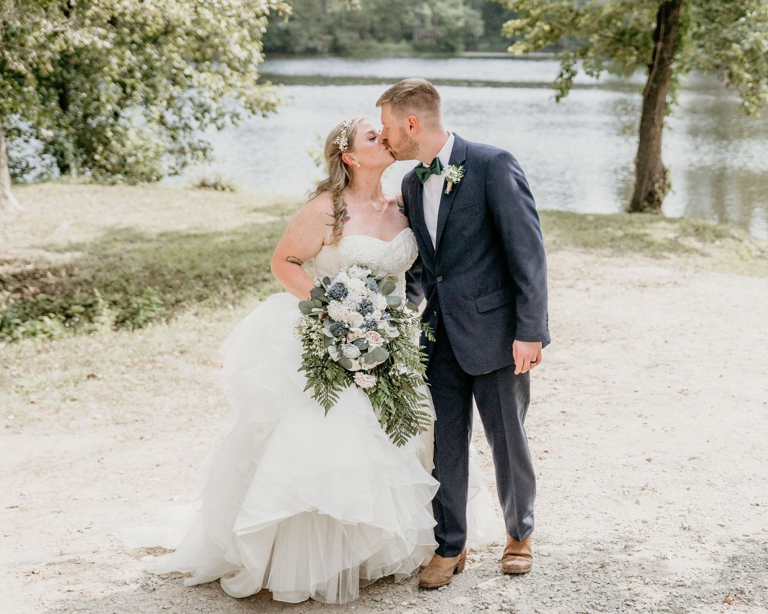 Romantic wedding photo with couple embracing by a serene lake wearing formal wedding attire and holding bouquet.