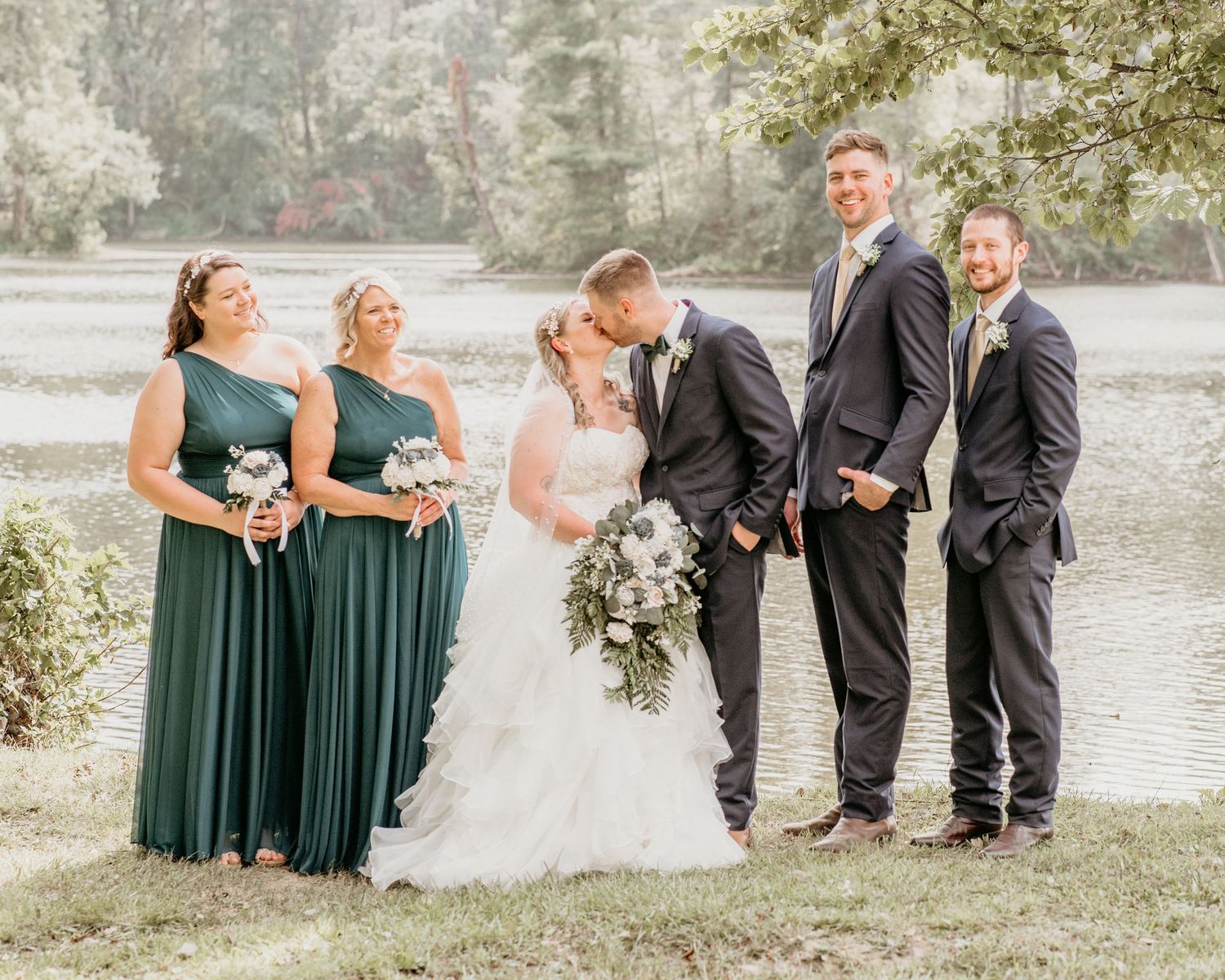 Wedding party in hunter green bridesmaid dresses and black suits poses together by a scenic lakeside.