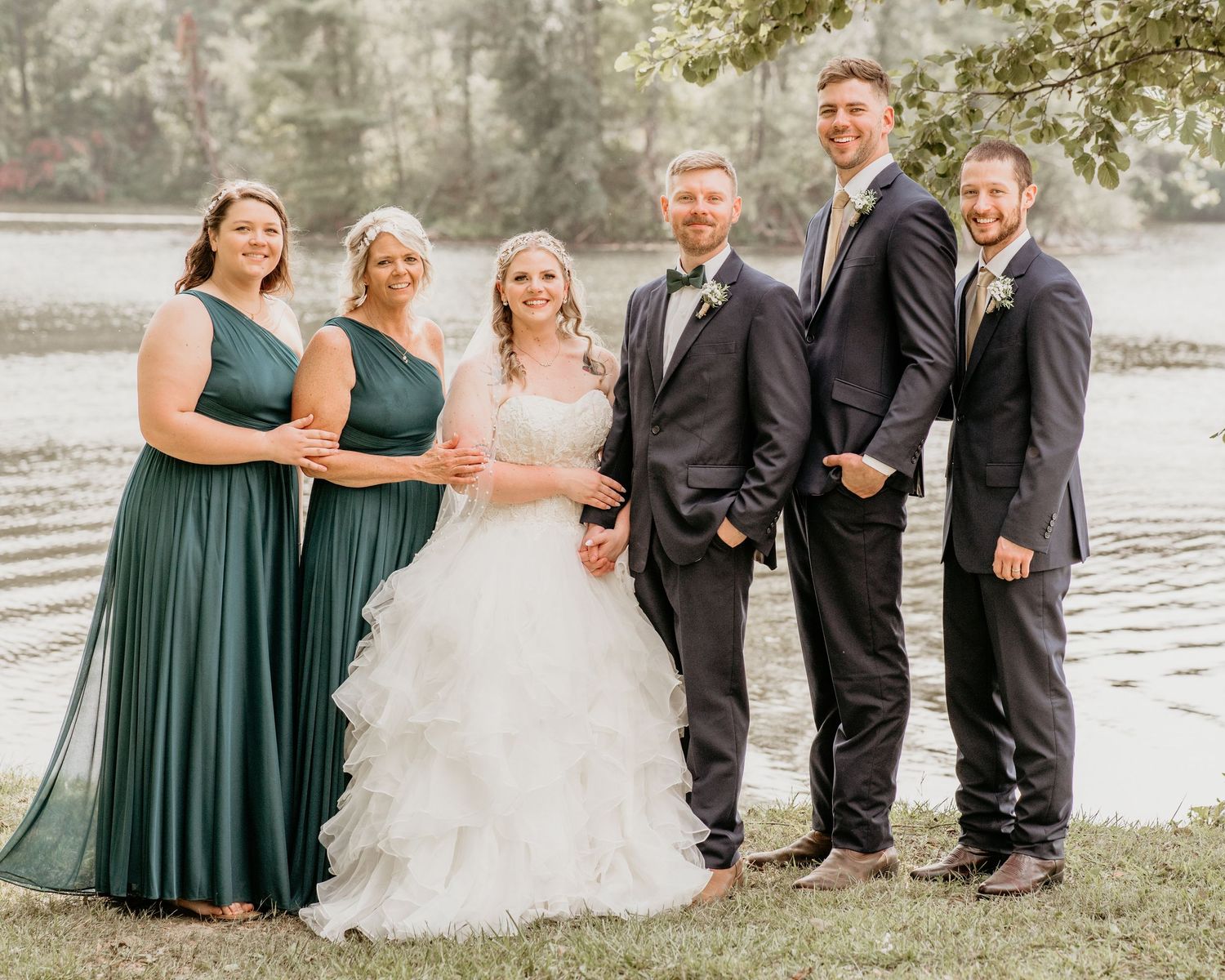Wedding party in dark green bridesmaid dresses and black suits pose by a serene lake for formal photos.