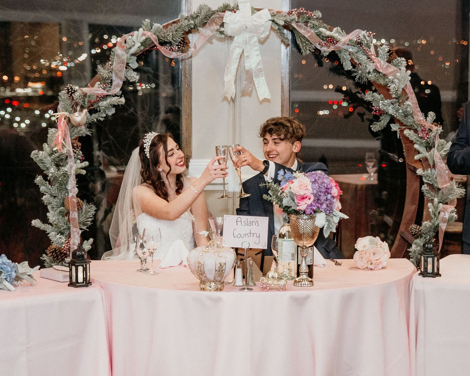 A wedding couple toasts at their sweetheart table under a floral arch with romantic lighting and pink decor.