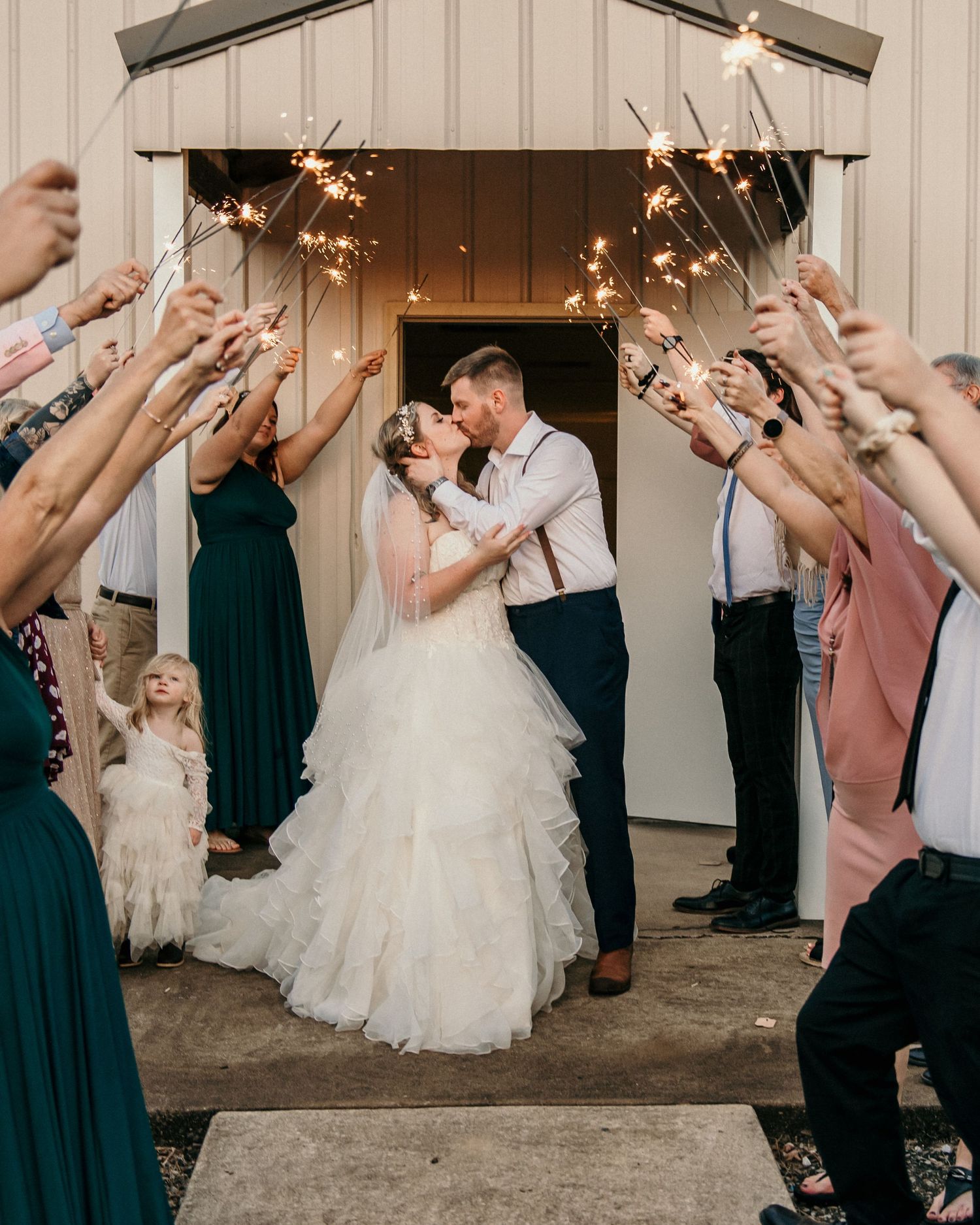 Wedding couple kissing under sparklers held by wedding guests in formal attire outside a white building entrance.