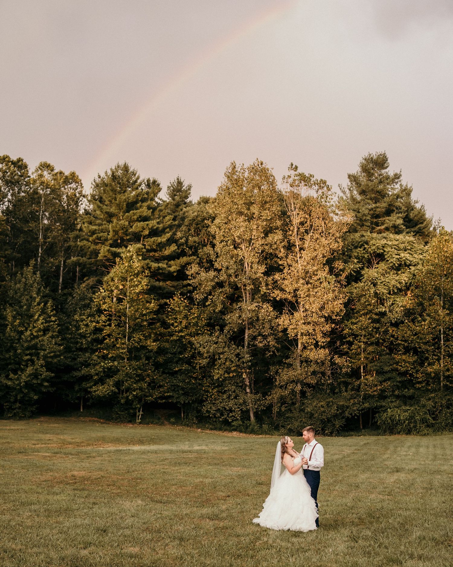 A couple embraces in a grassy field during golden hour with a rainbow and forest backdrop on their wedding day.