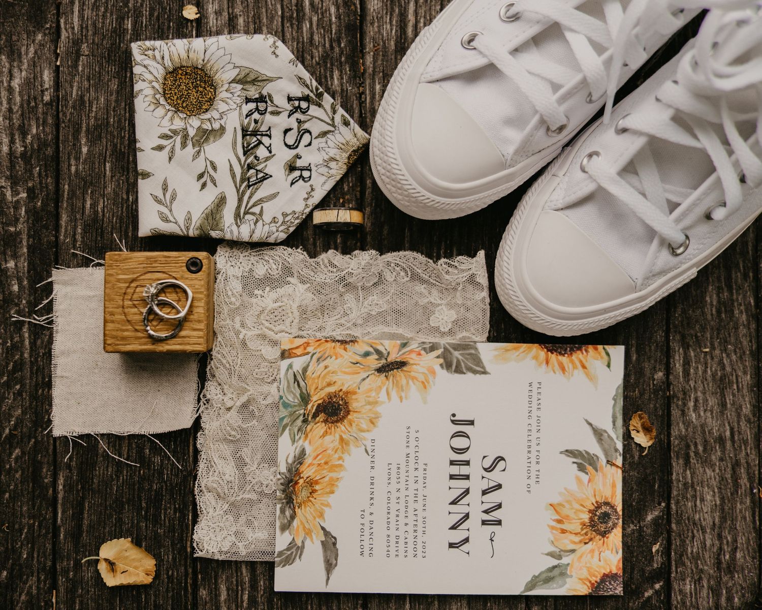 Wedding details flatlay with white sneakers, sunflower-themed invitation and wooden ring box on rustic surface.