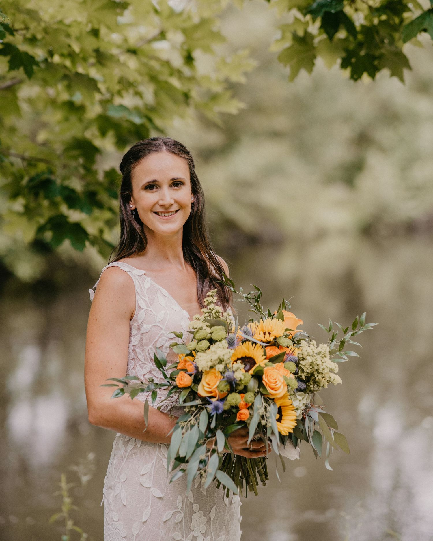 Bride in delicate lace dress holds vibrant orange and peach bouquet with eucalyptus against blurred green foliage background.
