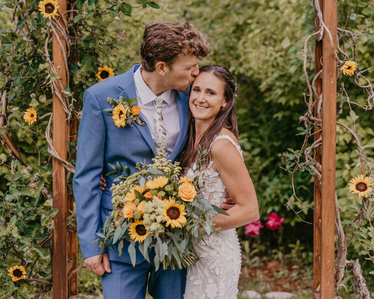 Rustic outdoor wedding ceremony featuring a couple posing under wooden arch decorated with bright sunflowers.