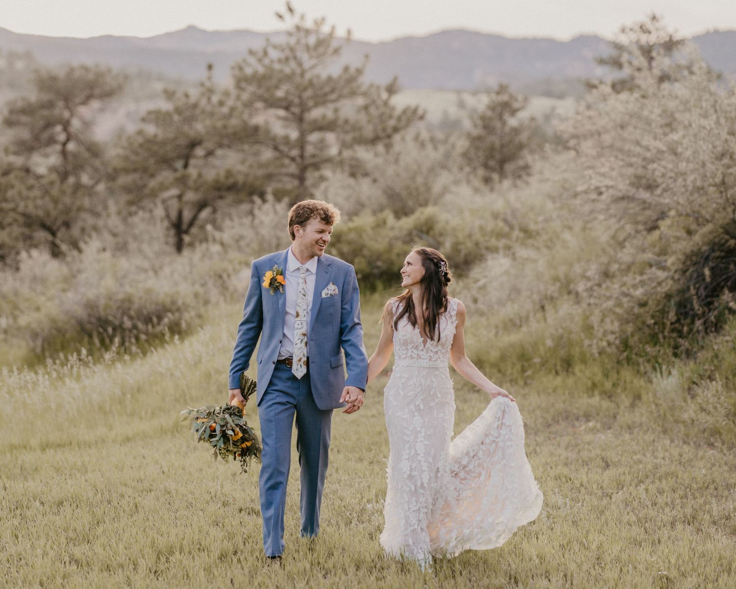 A couple walks hand in hand through a scenic mountain meadow at their wedding, wearing blue suit and lace white dress.