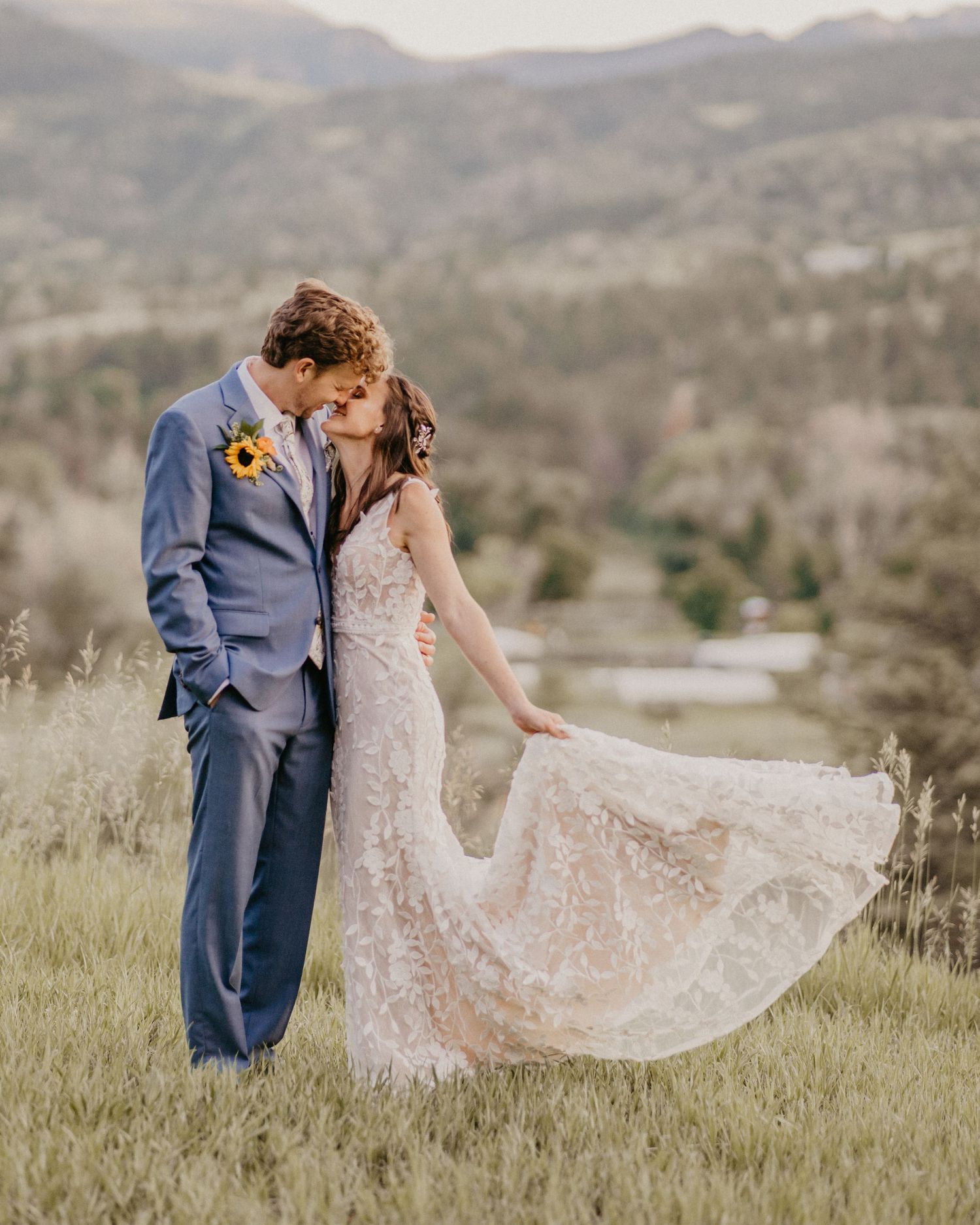 Romantic wedding couple embracing on grassy hilltop with mountains in background, bride's lace dress flowing in breeze.
