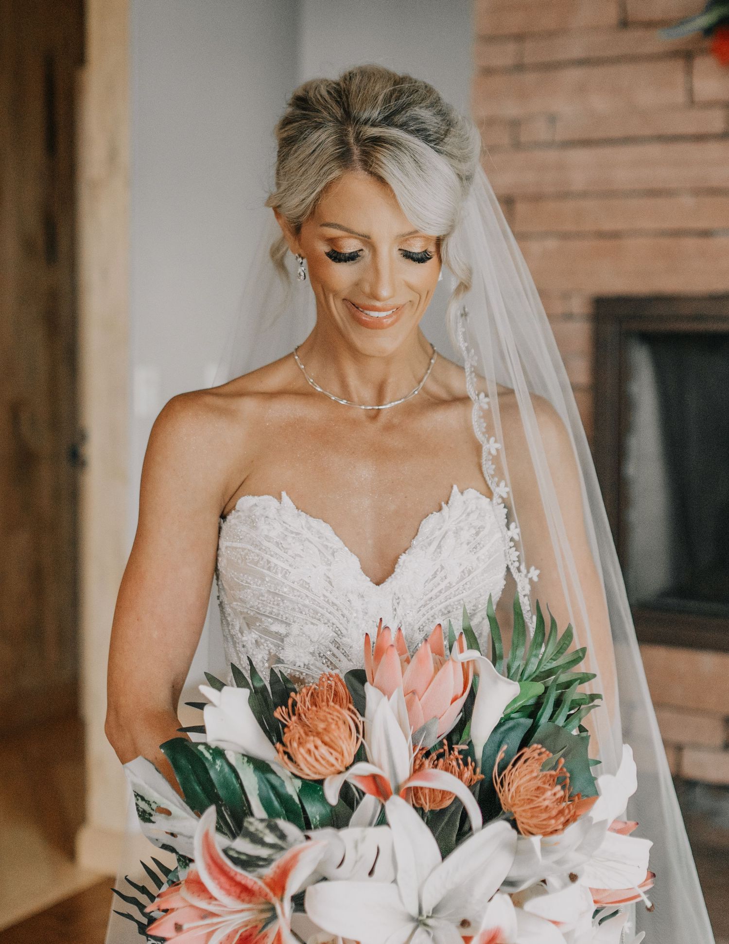 A bride in a strapless white lace dress holds a beautiful bouquet featuring pink proteas, peach roses and tropical greenery.