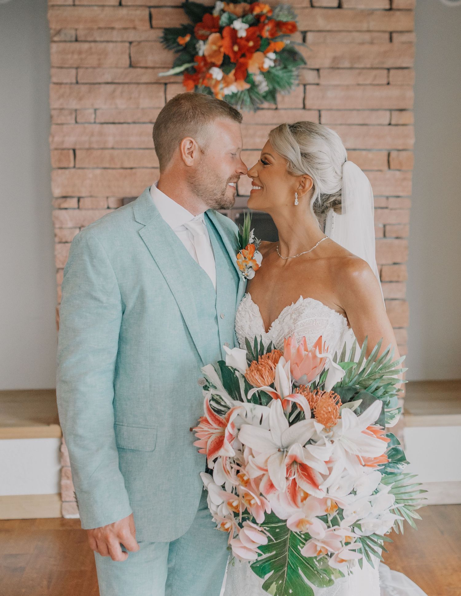 Wedding portrait with mint green suit and coral floral arrangements against brick wall.