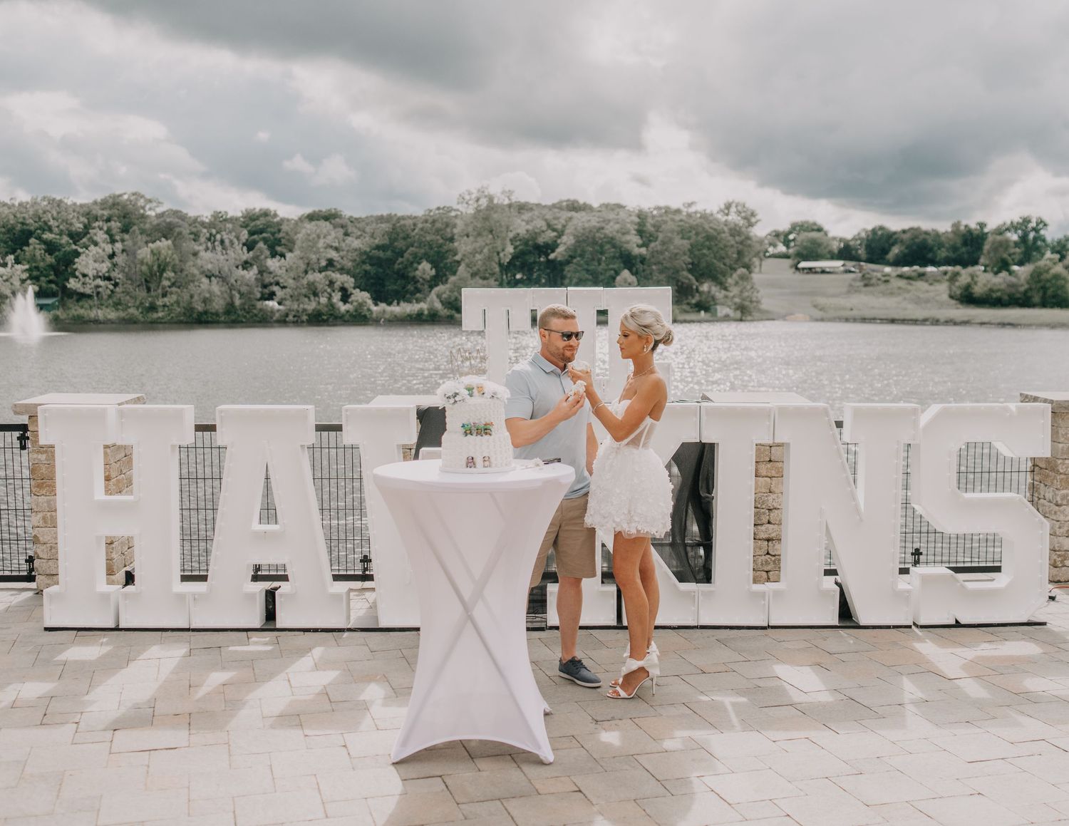 A couple in white outfits stands by a cocktail table on a deck with large white letters spelling custom last name behind them.