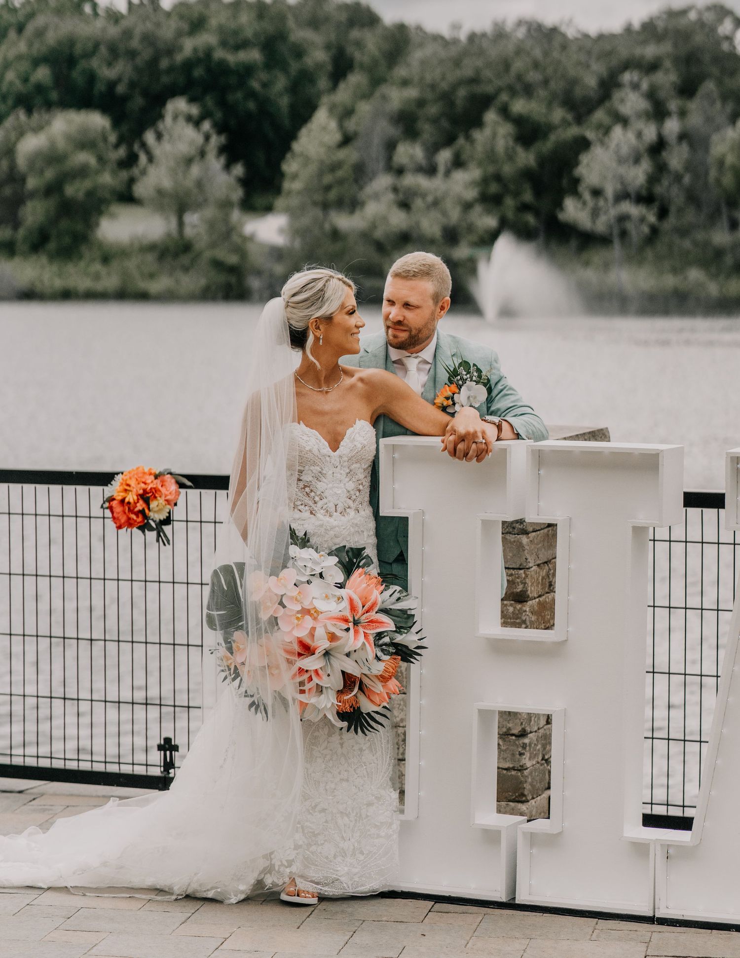 A bride in a lace gown and groom in gray suit share a romantic moment by a lake railing with coral floral bouquet. custom letters