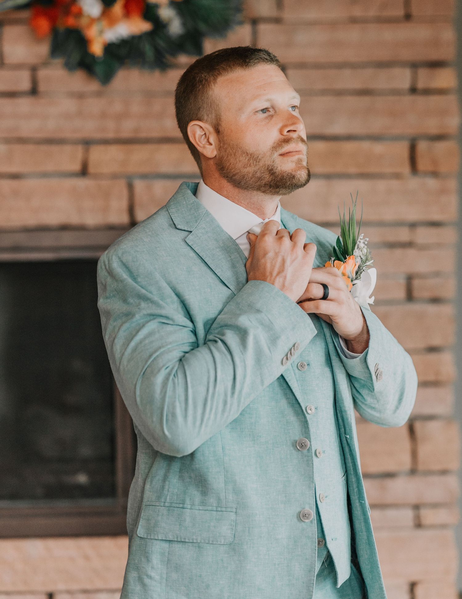 Elegant groom in mint green suit adjusting boutonniere against brick wall backdrop.