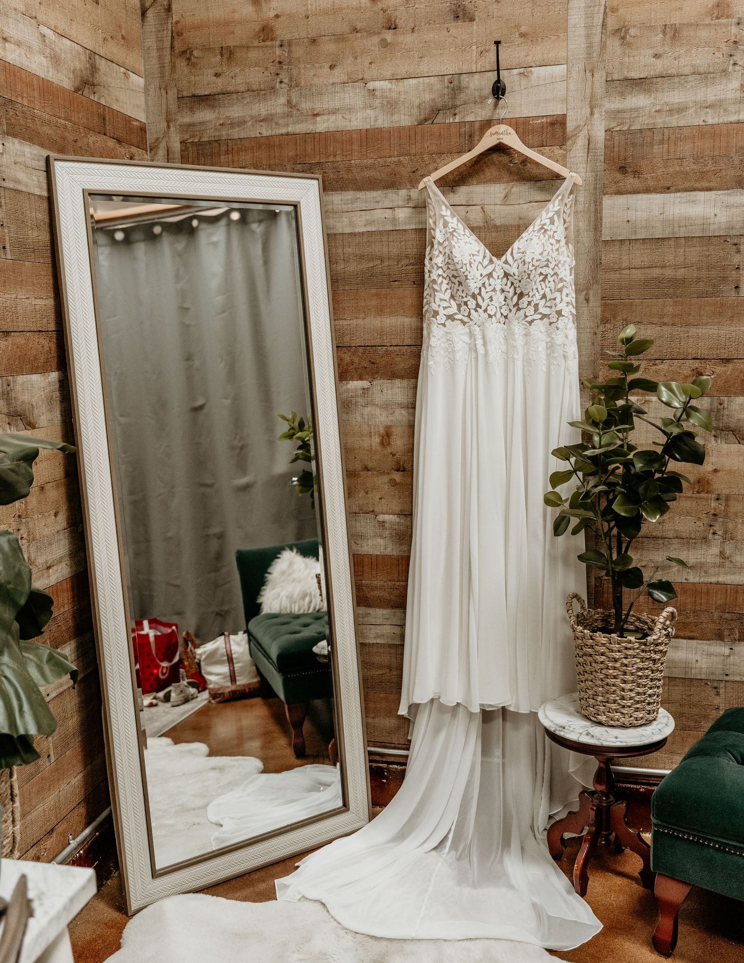 A flowing white lace wedding dress hangs next to a full-length mirror in a rustic wooden room with decorative greenery.