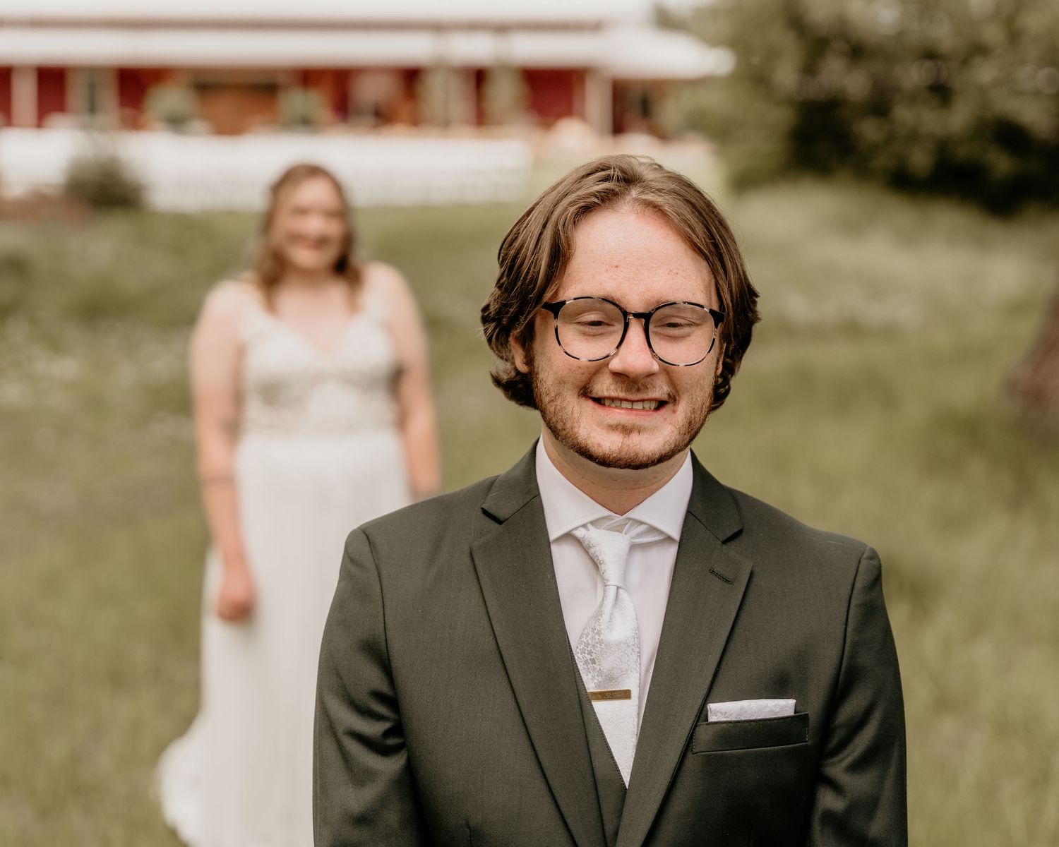 Wedding photo showing groom in dark suit and glasses with bride in white dress standing in background.